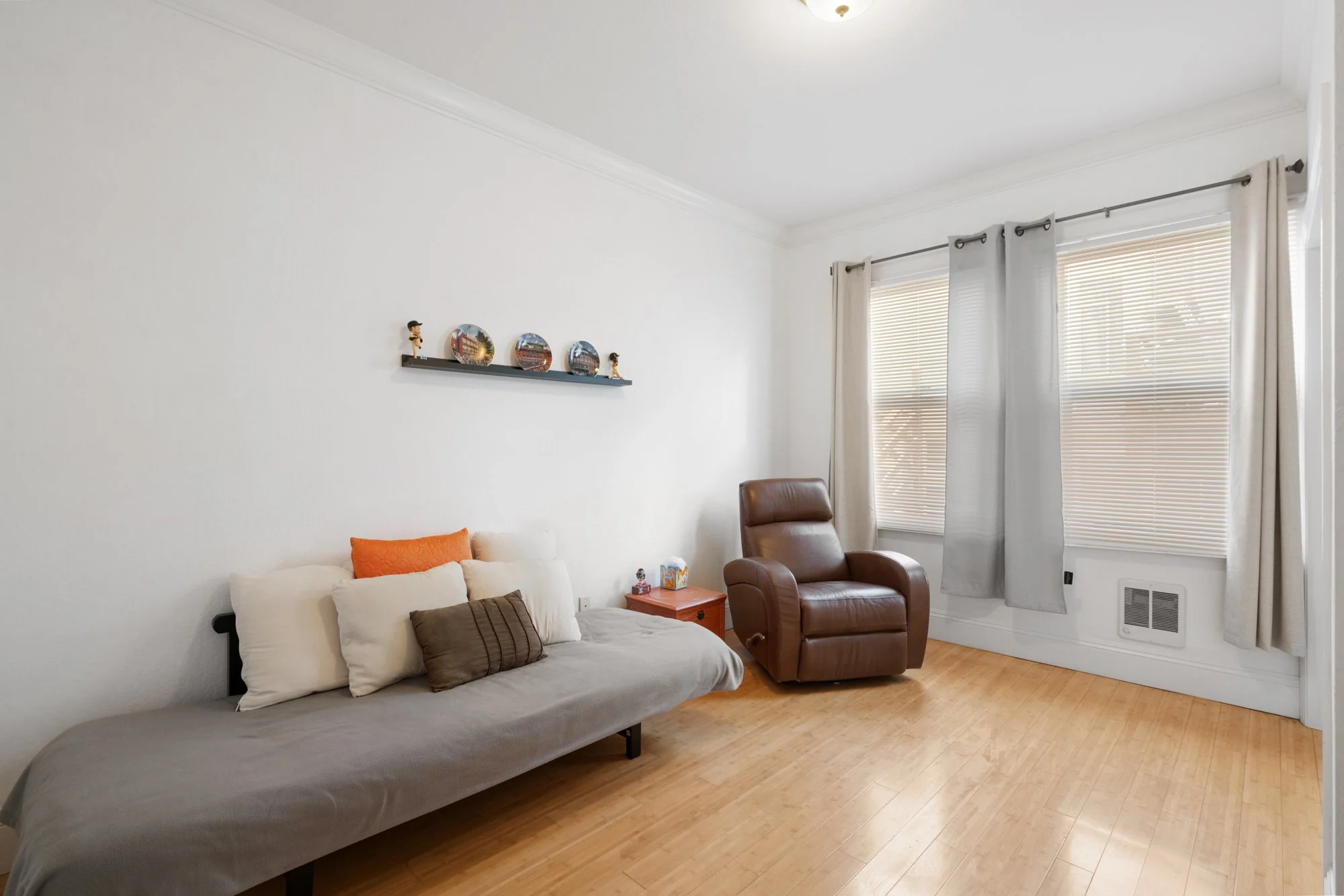 Living room with beige sofa, brown leather armchair, small orange side table, and window with blinds and curtains.
