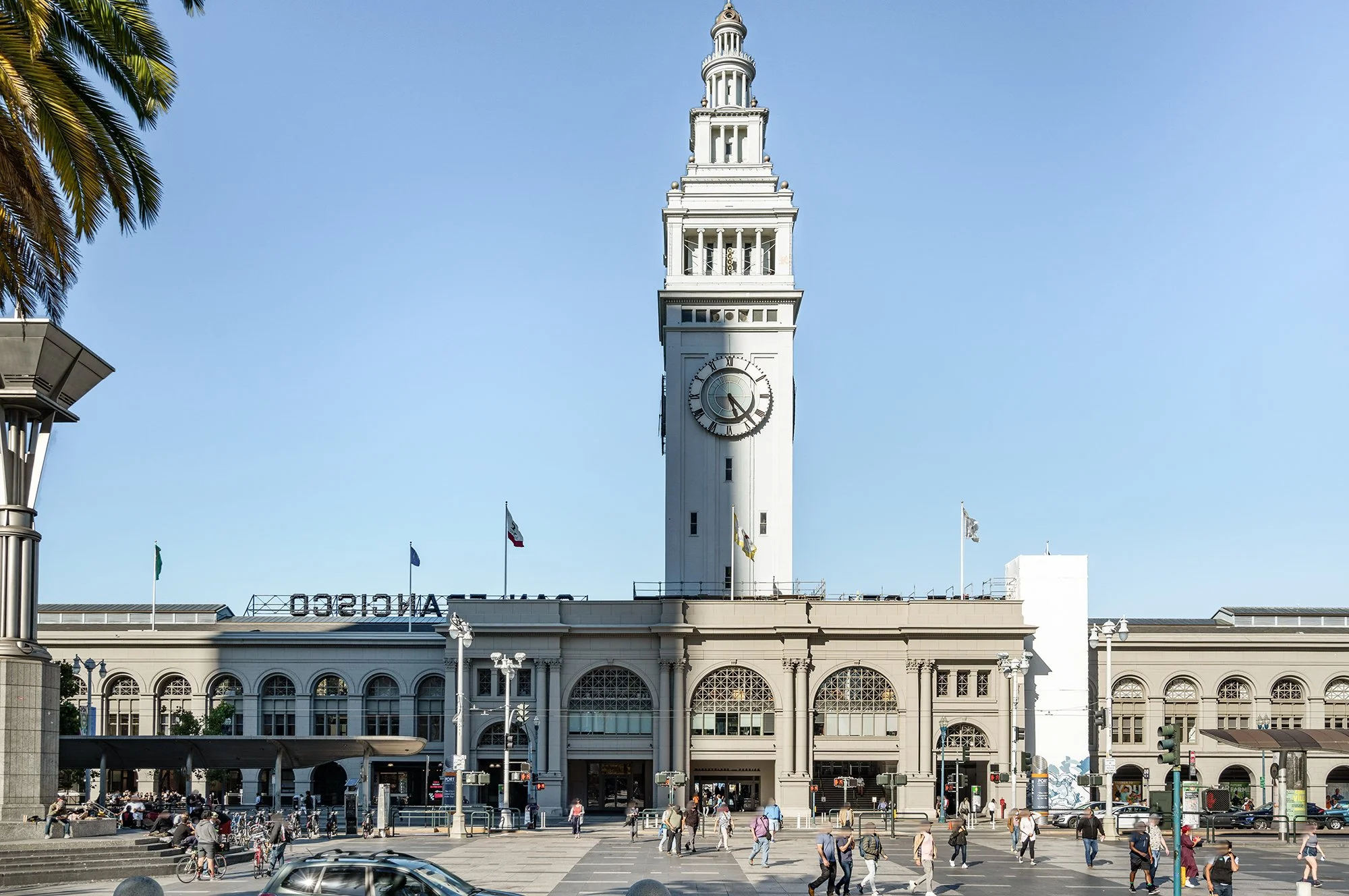 San Francisco Ferry Building with a prominent clock tower on a clear day, pedestrians walking in front, cars on the street, and flags flying.