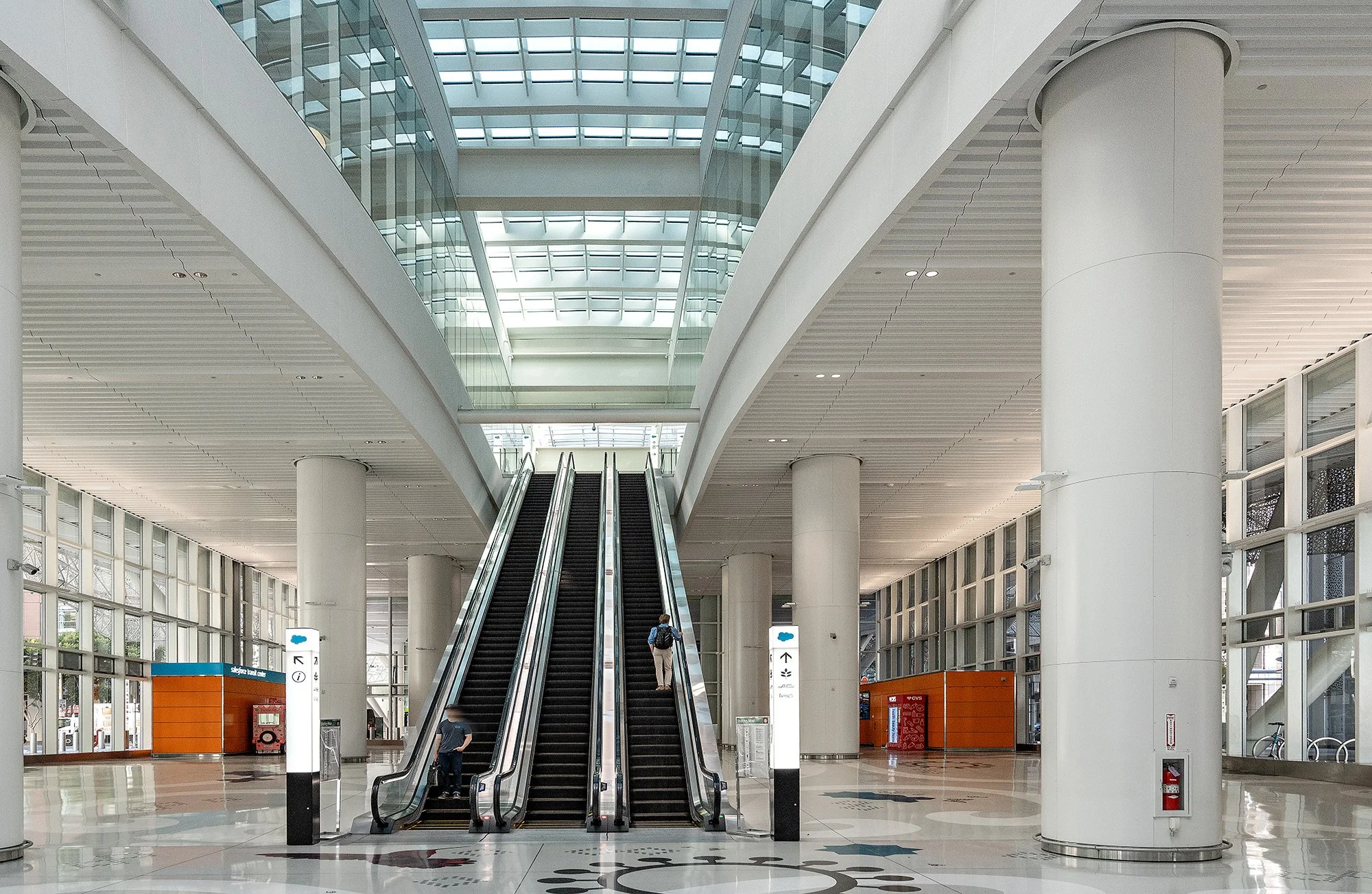 Inside a modern, spacious airport terminal with a high ceiling, large glass windows, and two moving escalators, with a few travelers ascending and descending.