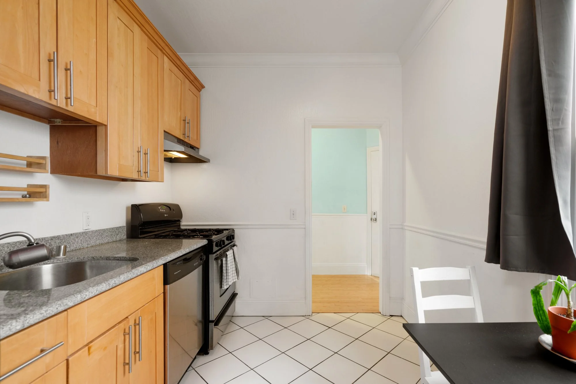Kitchen with wooden cabinets, gray countertop, stainless steel sink, black stove, dishwasher, tiled white floor, and a window with gray curtains.