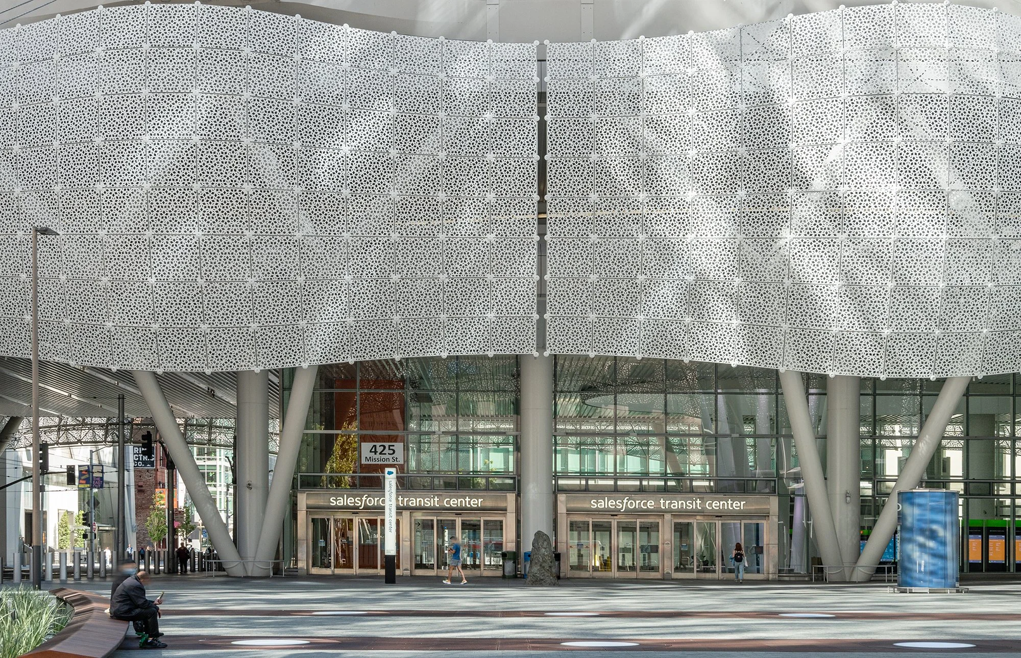 Exterior view of the Salesforce Transit Center in San Francisco with people walking and sitting outside, and a modern building with a decorative white facade.