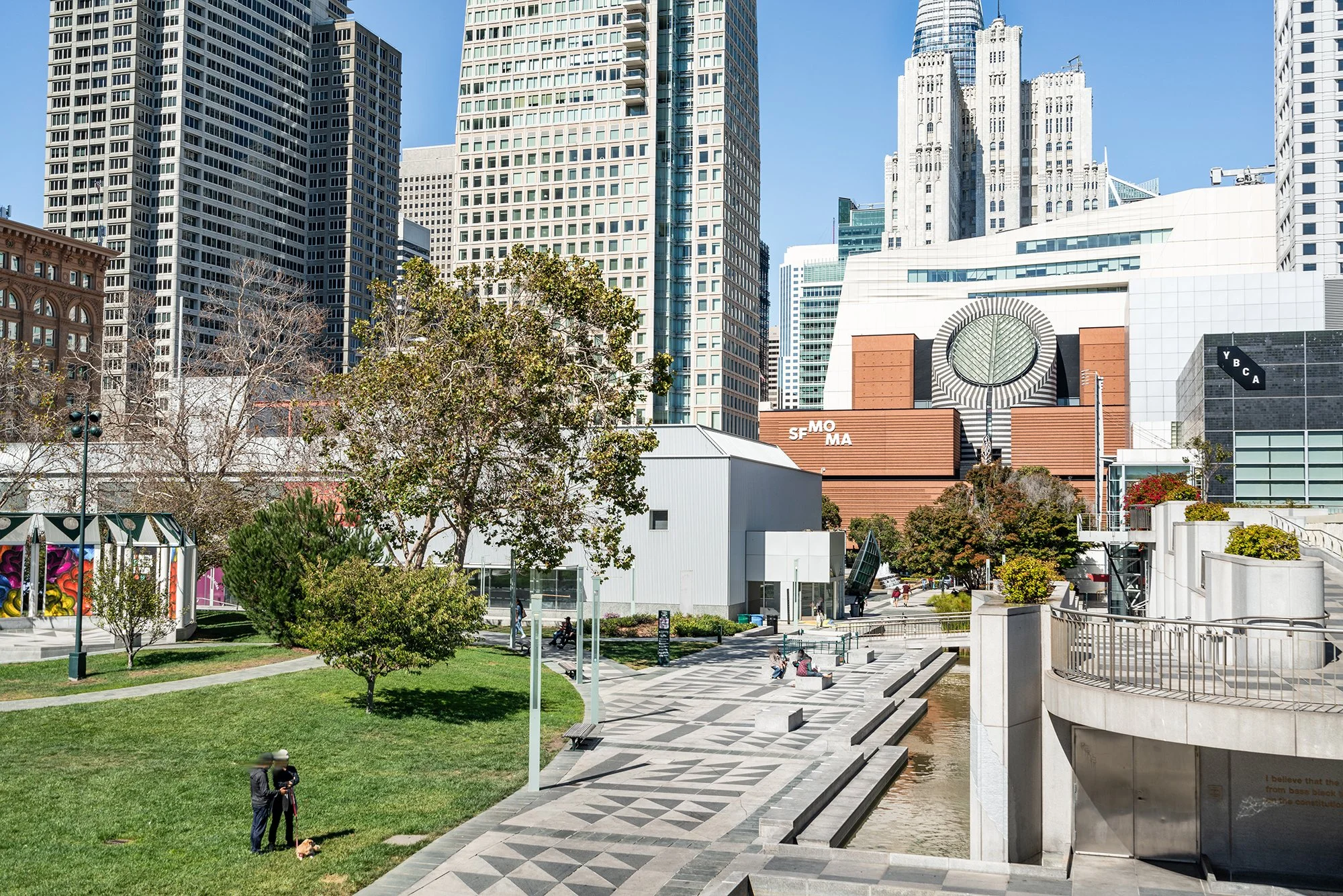 A city park with trees, grass, and a paved walkway along a water feature, surrounded by tall modern skyscrapers and buildings in downtown San Francisco on a clear day.