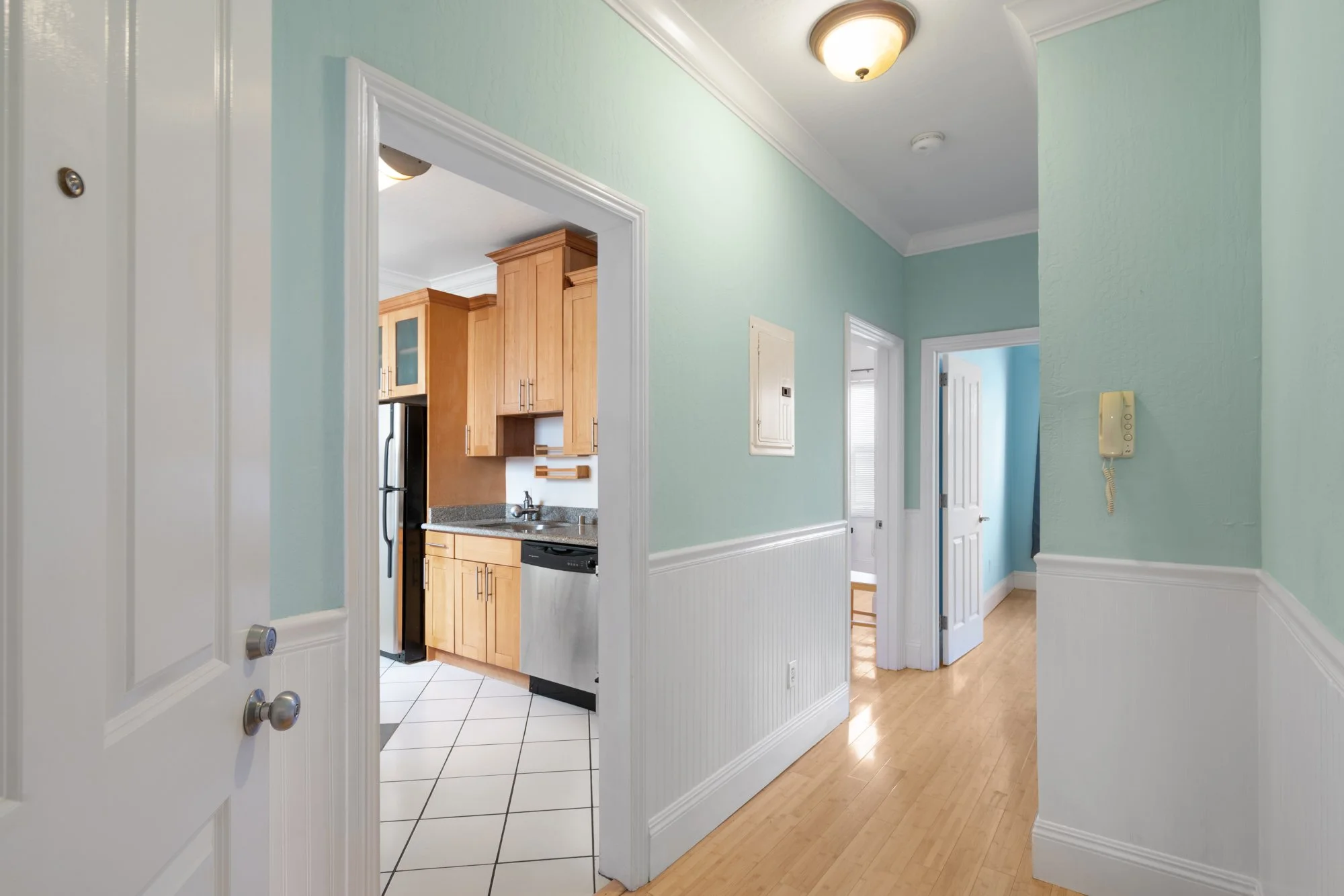 Interior view of a hallway with light green walls, white trim, and hardwood floors, leading to a kitchen with wooden cabinets and black appliances.