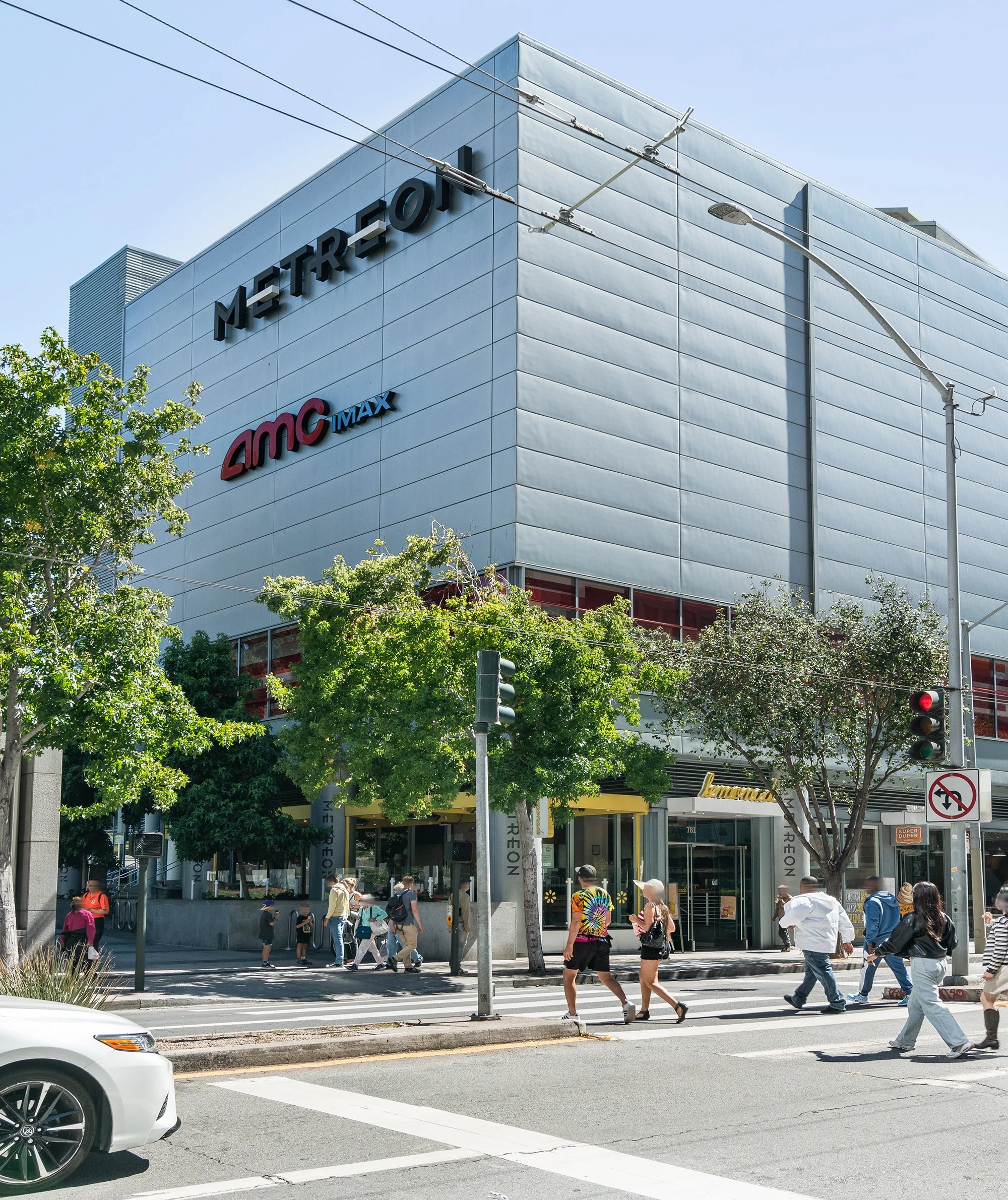 A busy city street scene in front of a large modern building with Metro and AMC IMAX signage. Pedestrians are crossing the street, and trees line the sidewalk.