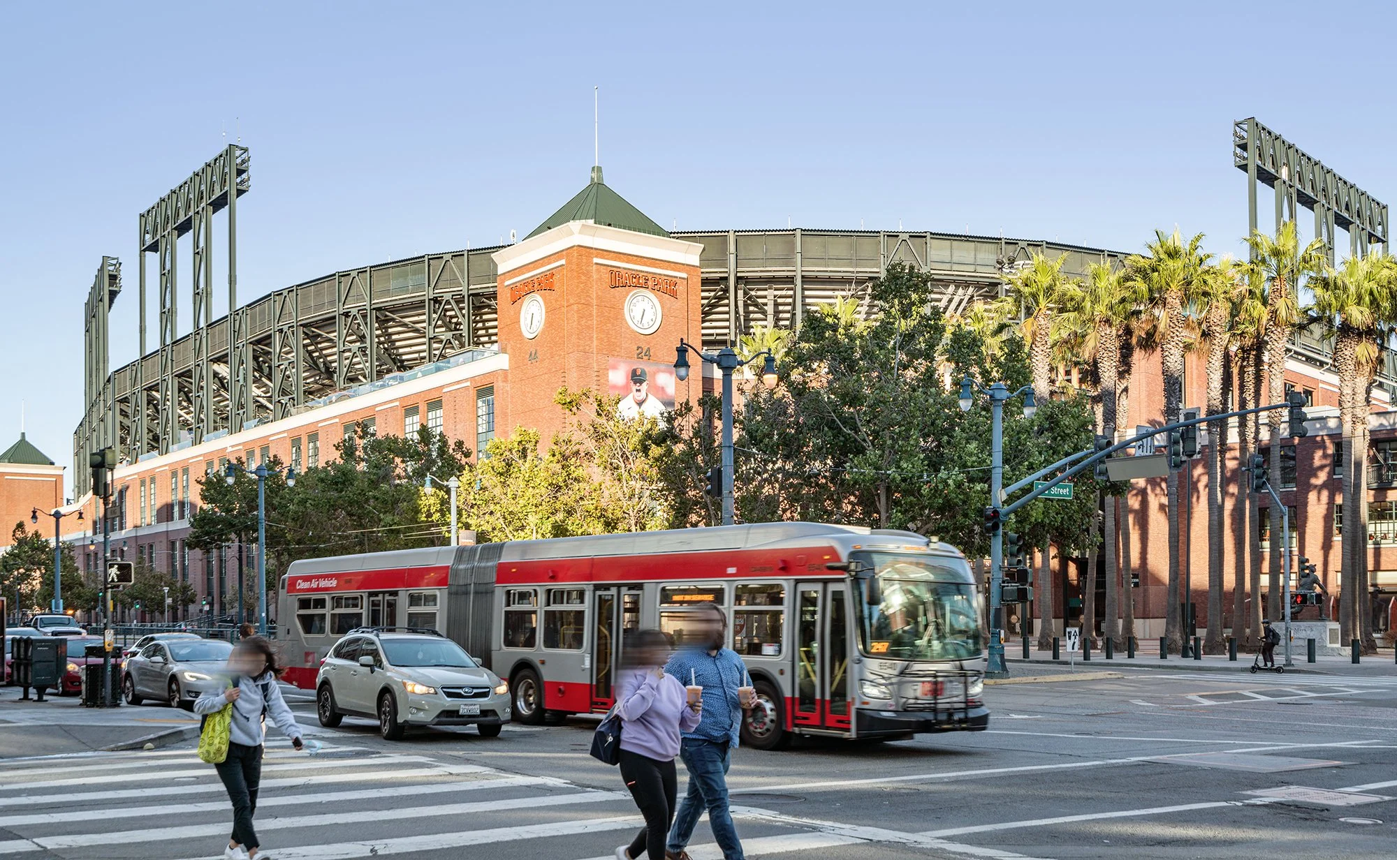 Street scene with pedestrians crossing at crosswalk, approaching a red and white bus, and Oracle Park stadium with palm trees in the background.
