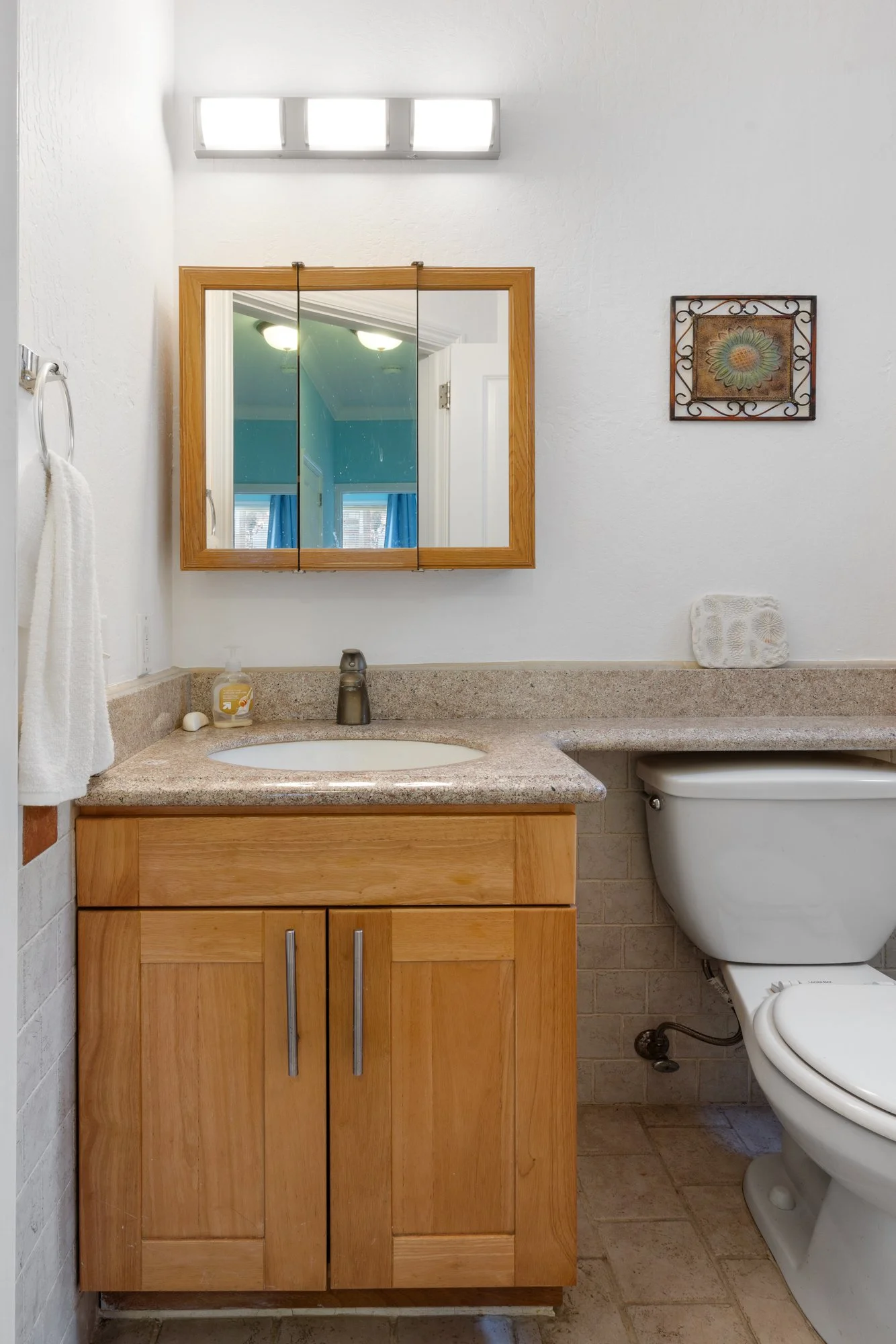 Bathroom with a wooden cabinet, sink, mirror, toilet, and wall decor, with a soap dispenser and a towel hanger.