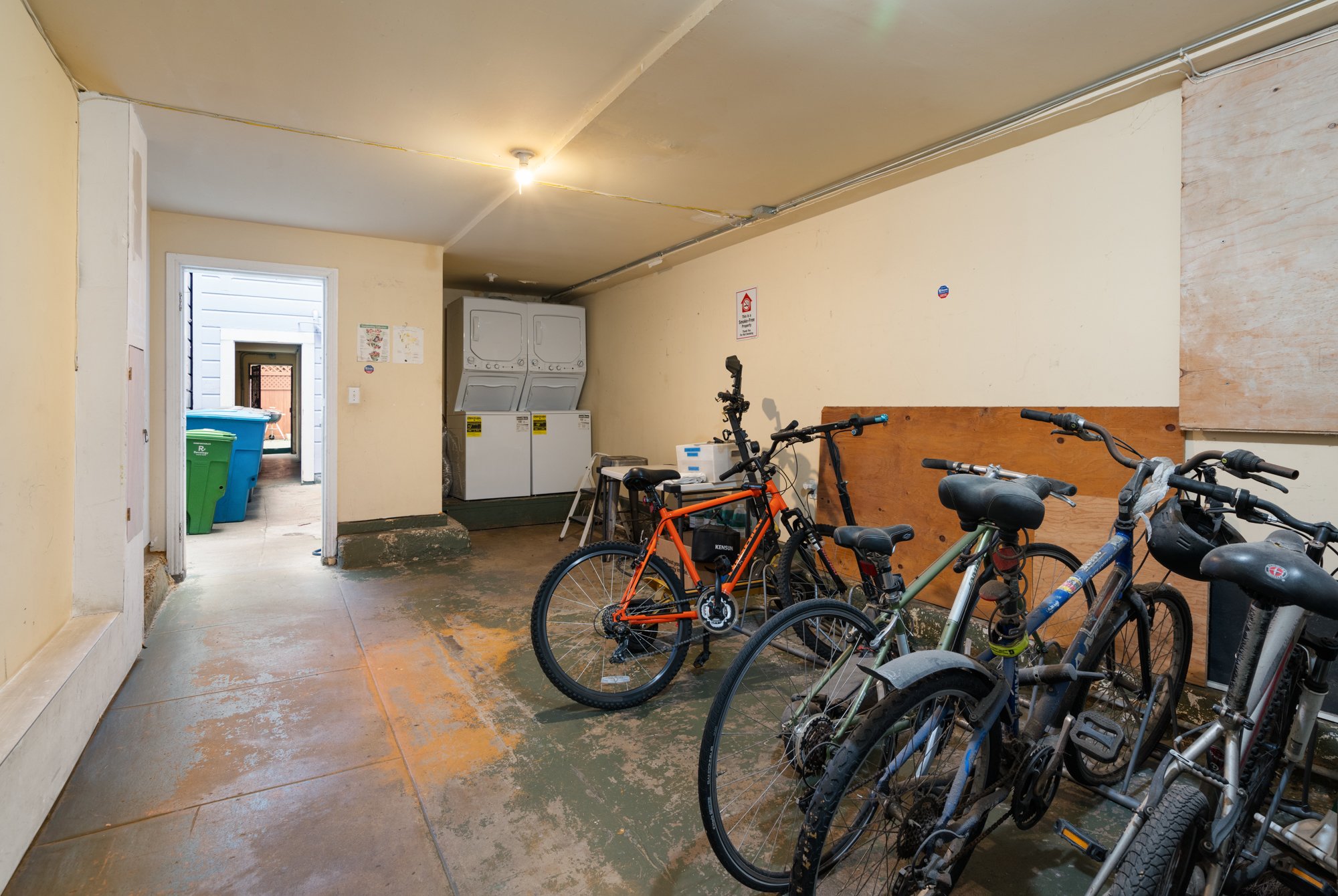 Indoor laundry and bike storage room with washing machines, bicycles, and two trash bins visible through an open door.