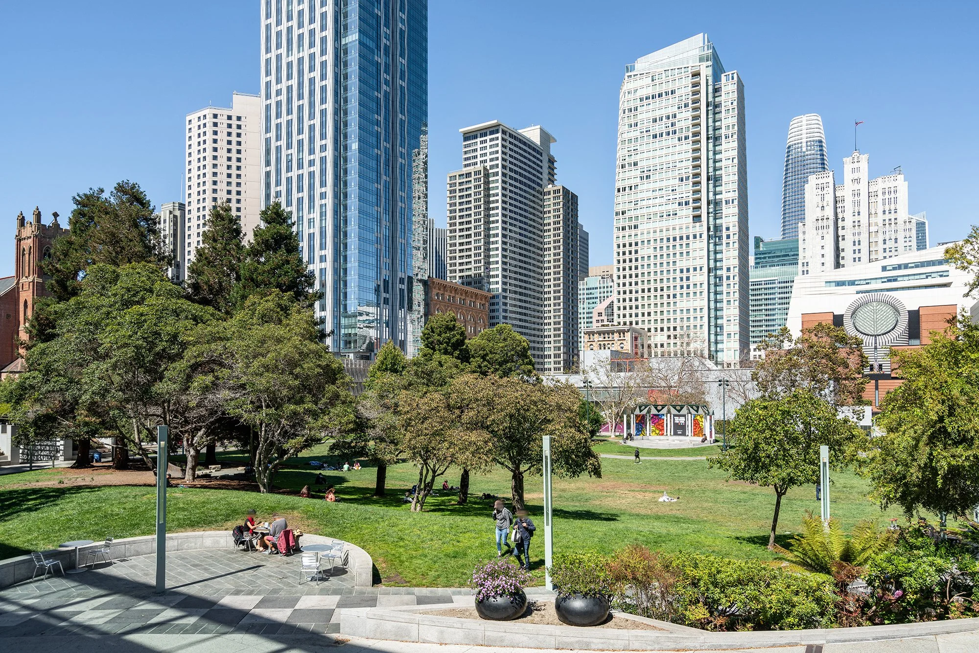 View of a city park with green trees, people sitting and walking, surrounded by tall skyscrapers in the background on a sunny day.
