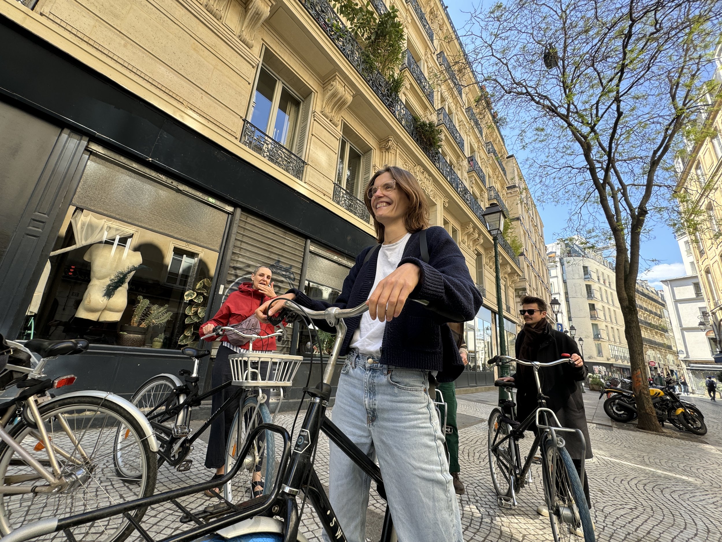 Vrouw met brillen en een zwarte jas, die op een fiets zit op een drukke stadsstreet met historische gebouwen, mensen en fietsen.