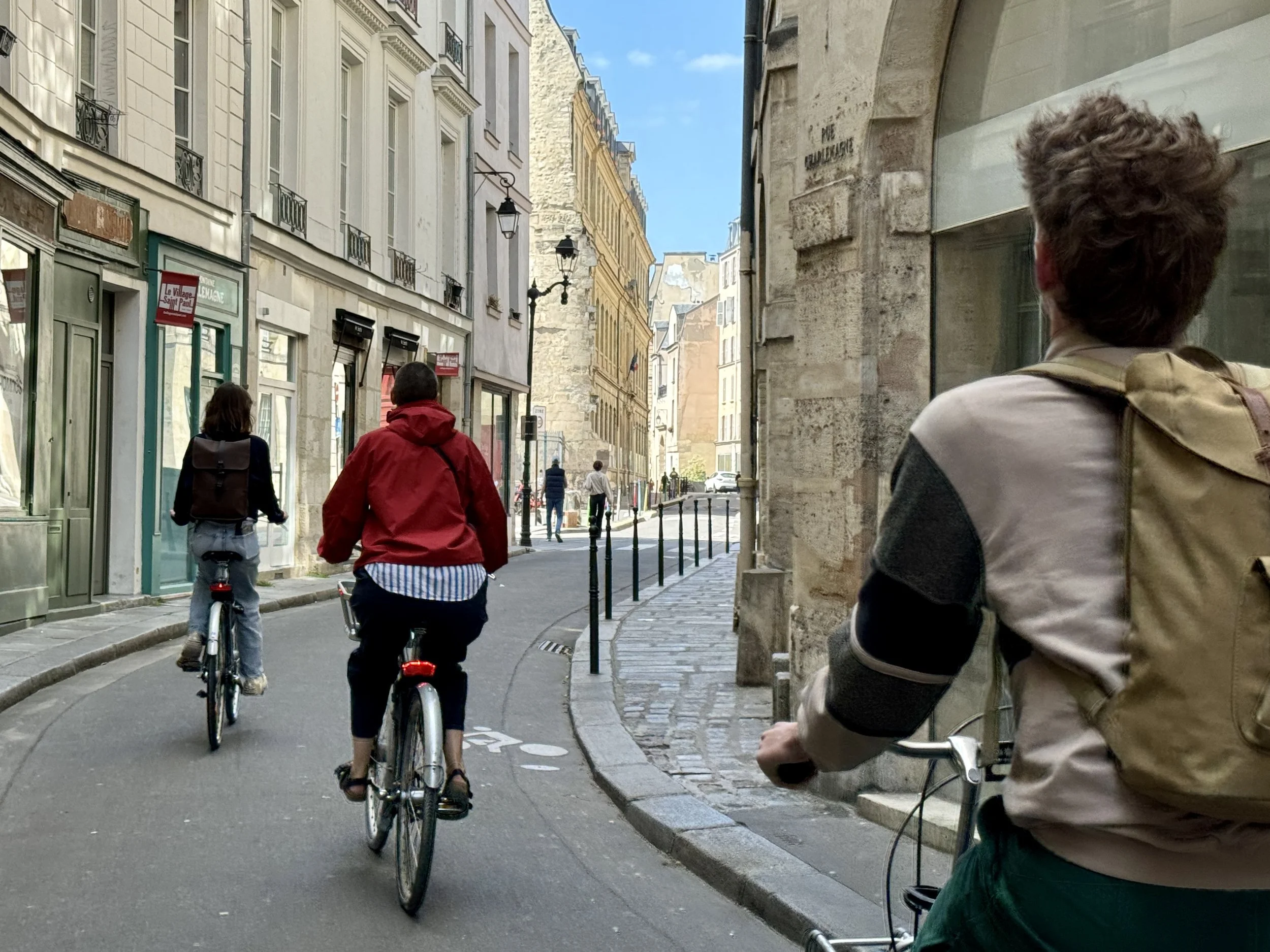 Drie mensen fietsen door een smalle straat met historische gebouwen in Parijs, met een blauwe hemel en een zonnetje. Bike tour in Paris. Must see things in Paris. Slow travel in Paris