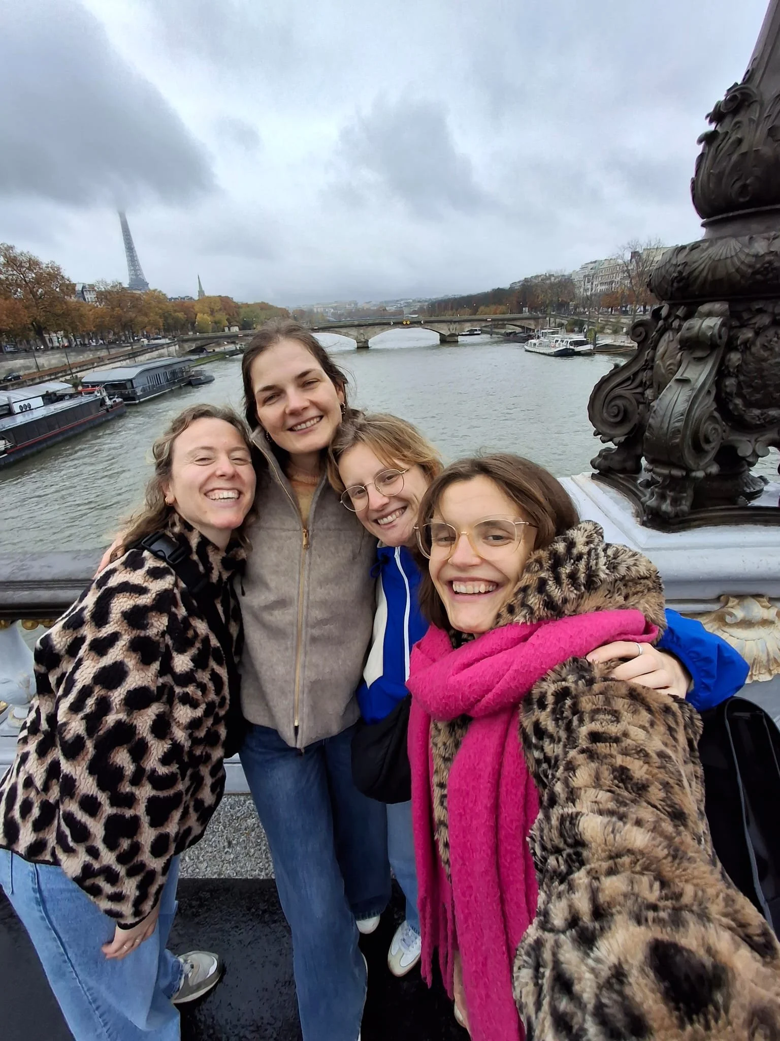 Vijf vrouwen die samen aan de waterkant in Parijs staan, met de Eiffeltoren op de achtergrond en een brug over de rivier. Ze lachen en lijken plezier te hebben. Wandeltocht. Nederlandse gids in Parijs