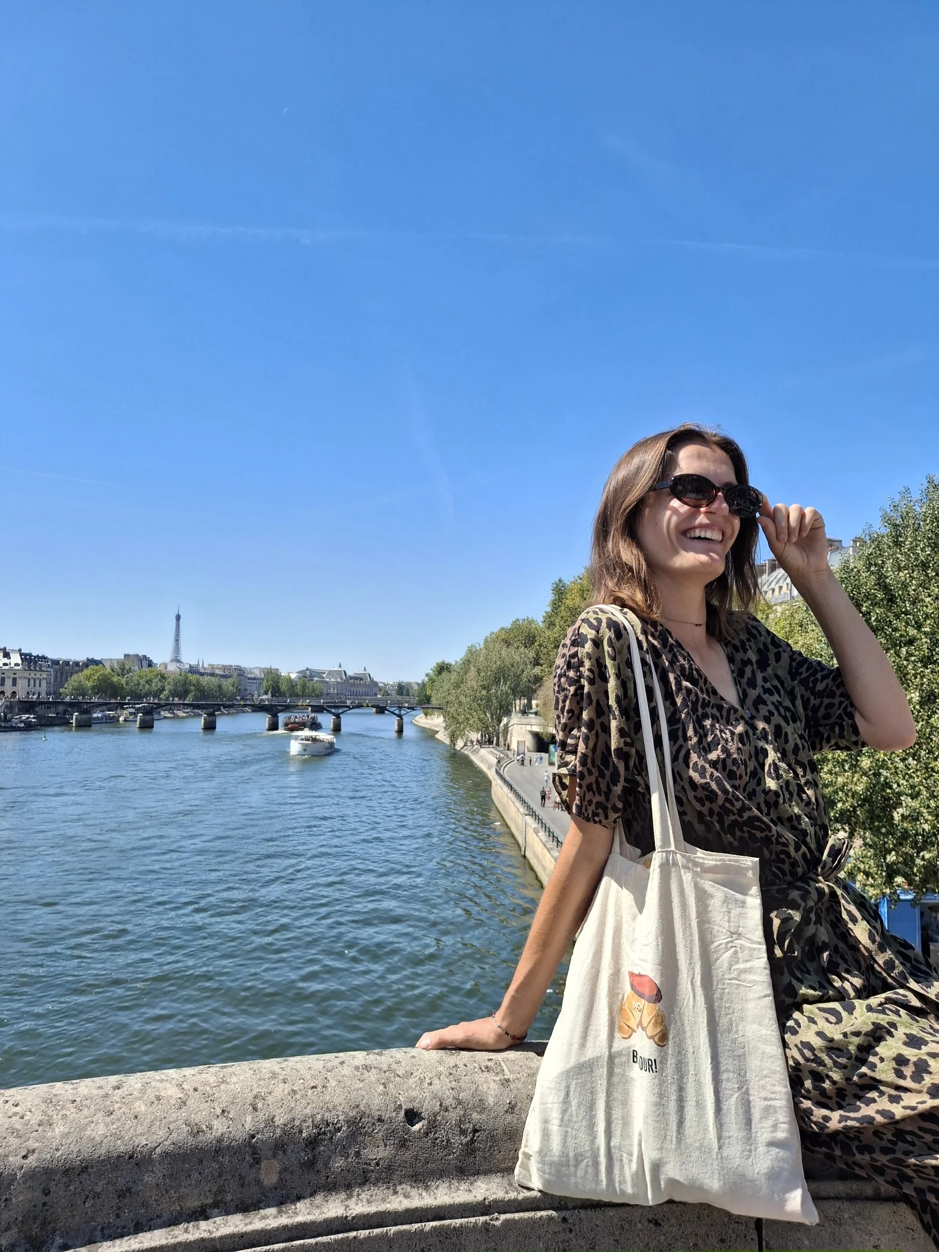 Nederlandstalige gids in Parijs, lachend en poserend naast de Seine-rivier in Parijs, met de Eiffeltoren op de achtergrond op een zonnige dag.