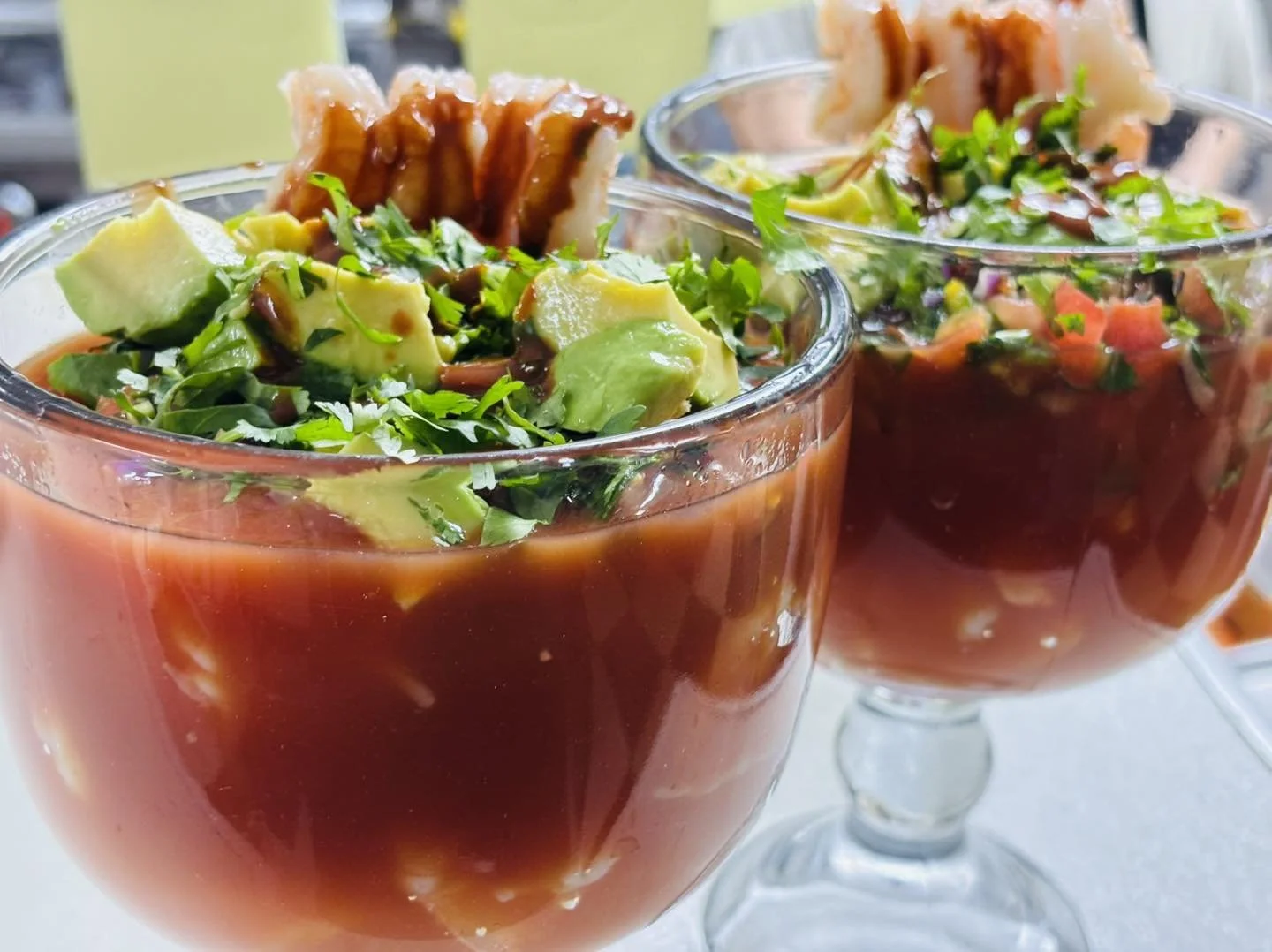 Two glasses of Mexican tomato cocktail with chopped vegetables, avocado, cilantro, and chili, garnished with pork rinds.