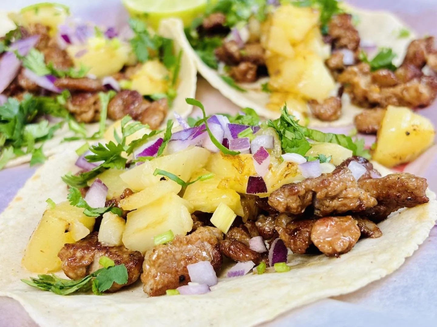 Three tacos filled with seasoned meat, pineapple chunks, chopped onions, cilantro, and green onions on soft tortillas.
