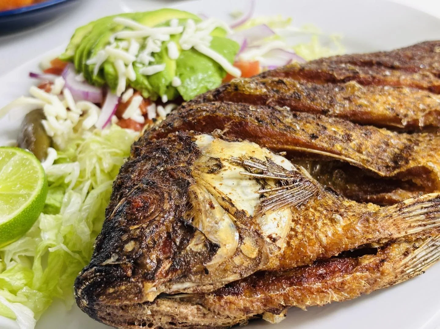 Fried whole fish with crispy skin served with a side of fresh salad, lime, shredded cheese, and vegetables.