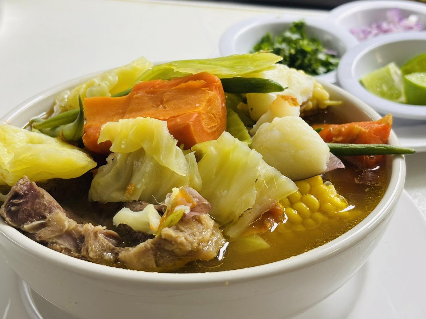 A bowl of vegetable soup with carrots, potatoes, corn, lettuce, and other vegetables, with side dishes of lime wedges and chopped herbs.