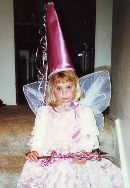A young girl dressed as a fairy princess with pink wings, a pink dress, and a tall pink cone hat, sitting indoors.