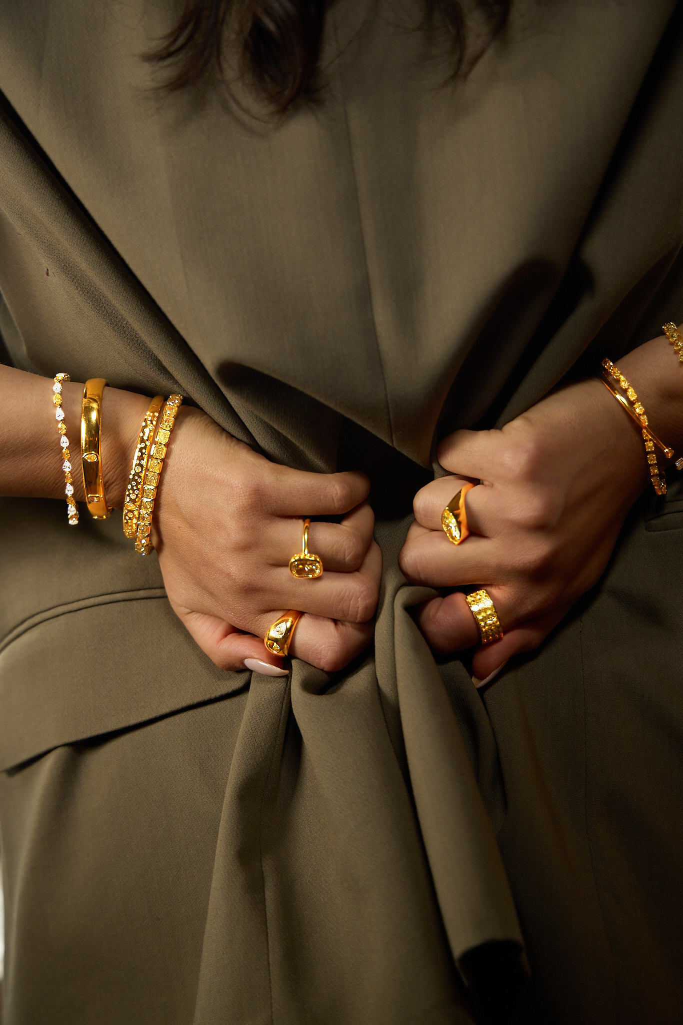 Close-up of a person's hands with multiple gold jewelry bracelets and rings, wearing an olive-green blazer.