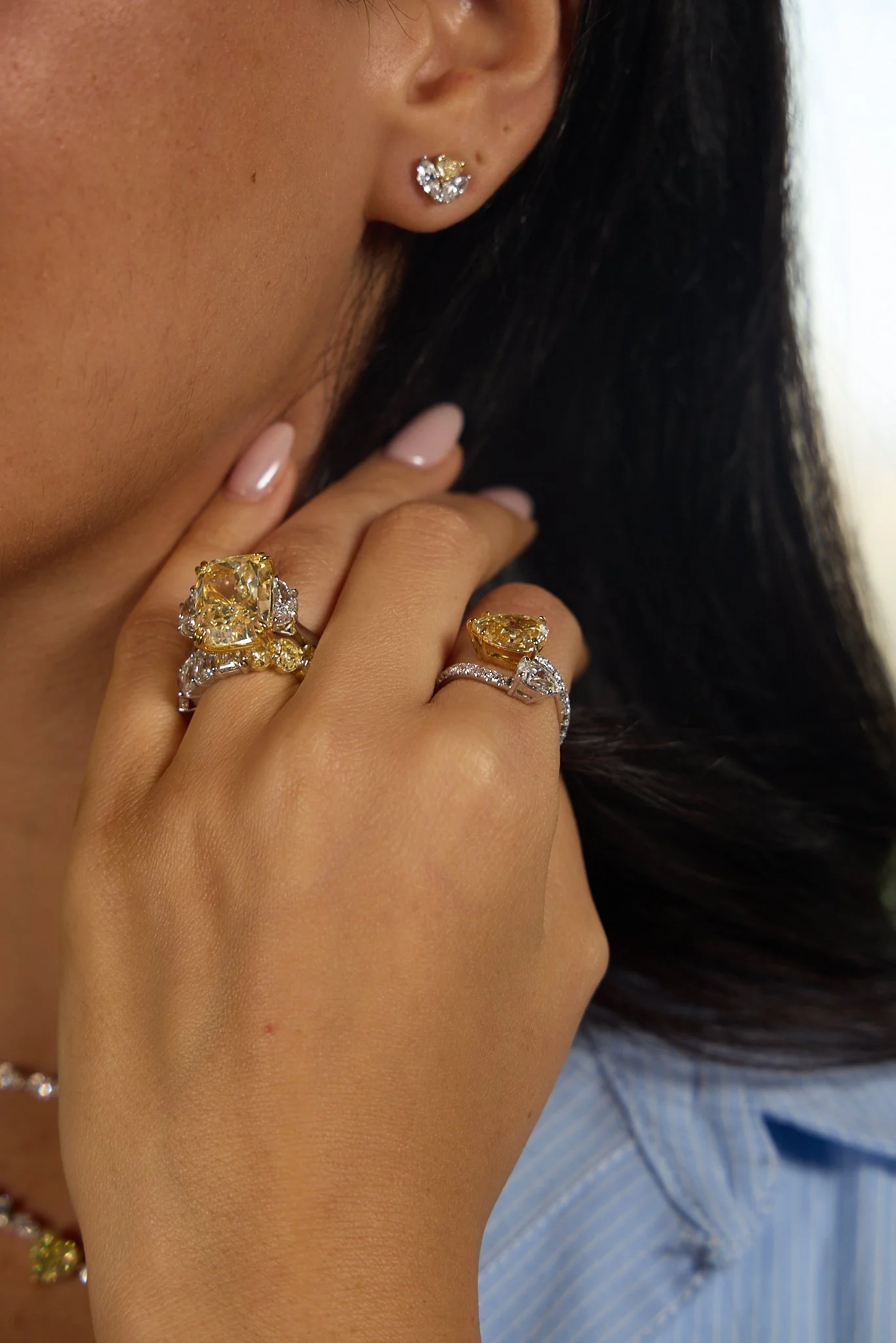 Close-up of a woman's face and hand, showing multiple rings with large yellow and clear gemstones, and an earring with a cluster of clear gemstones.