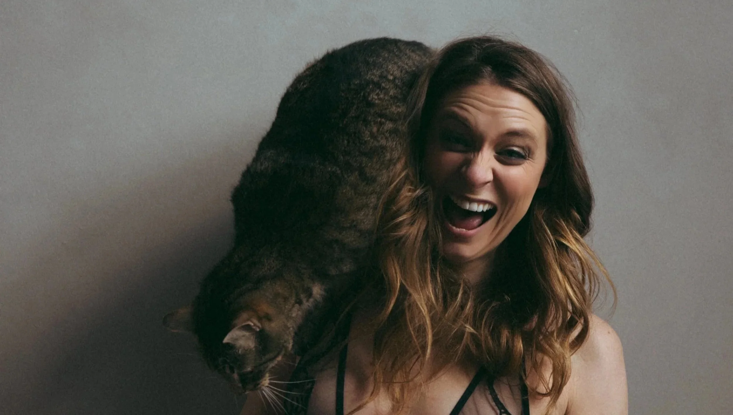 A woman with wavy brown hair smiling as a large tabby cat rests on her shoulder.