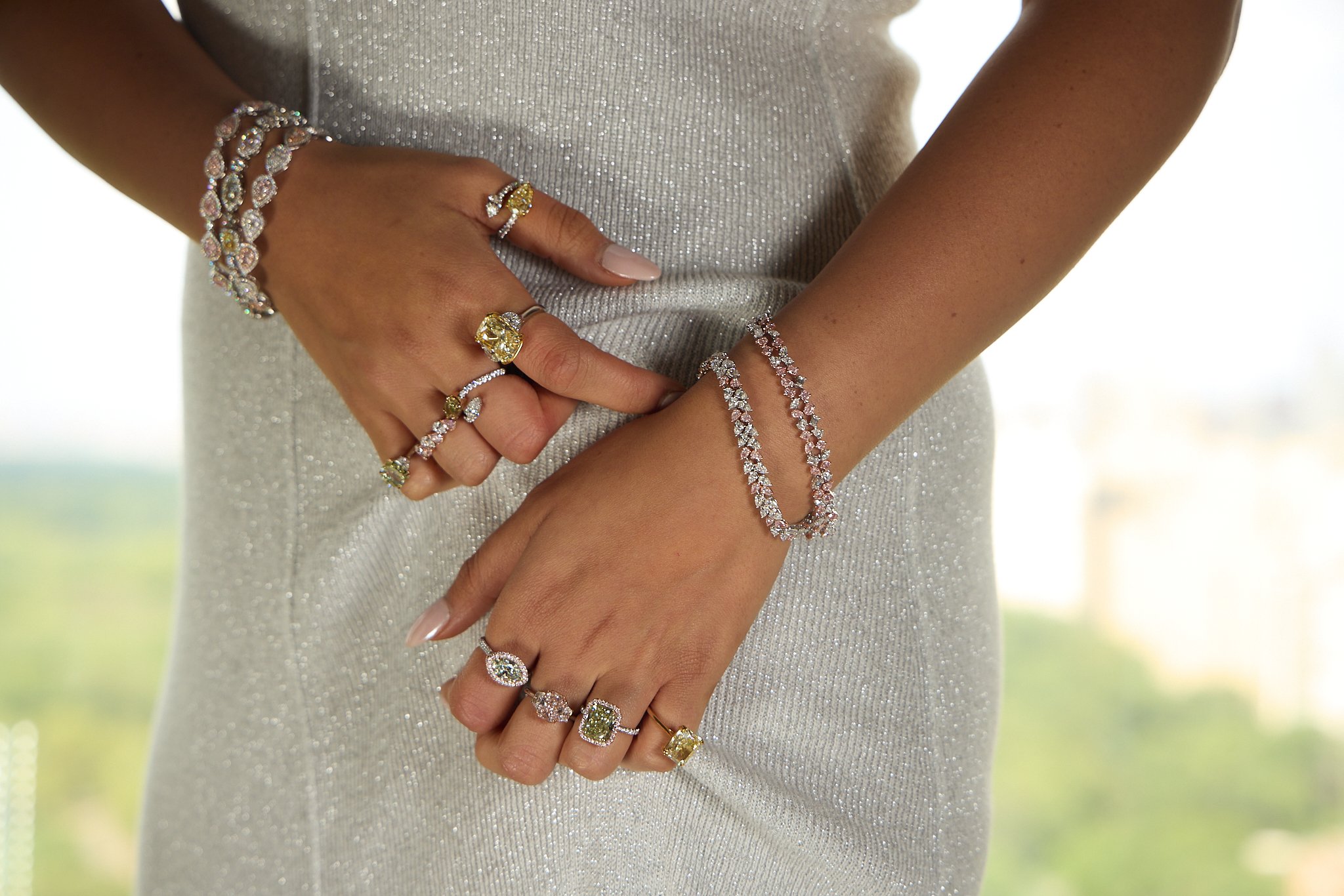 Close-up of a woman's hands and arms adorned with multiple gold and diamond rings, bracelets, and a pearl bracelet, in front of a shimmering silver dress.
