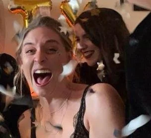 Two women celebrating with confetti, one wearing balloon animal-shaped headgear, at a festive event.