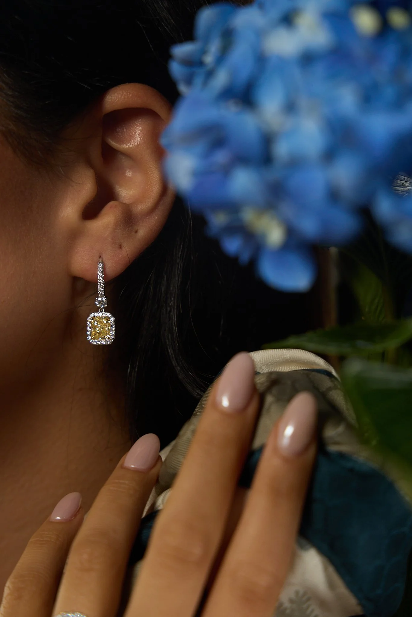 Close-up of a woman’s ear wearing a diamond and yellow gemstone rectangular drop earring, with hand and manicured nails near a blue flower.