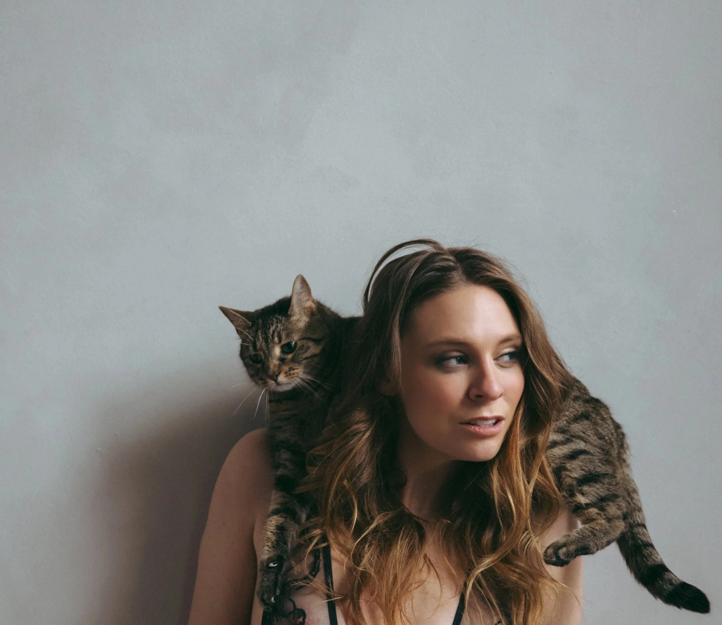 A woman with long wavy brown hair and a tabby cat draped over her shoulders, both positioned against a plain gray wall.