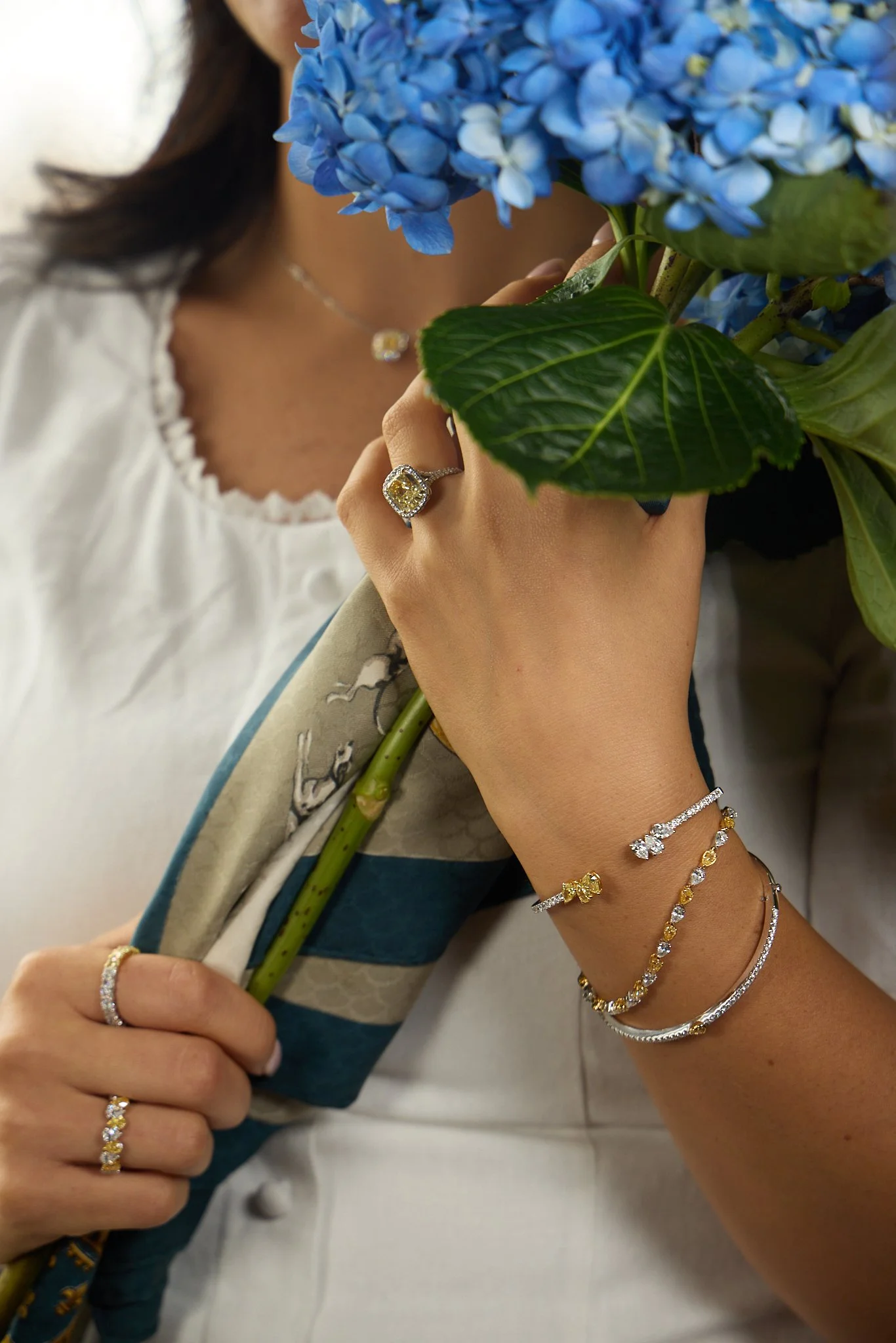 A woman holding a bouquet of blue hydrangeas, wearing diamond and gold jewelry including rings, bracelets, and a necklace.