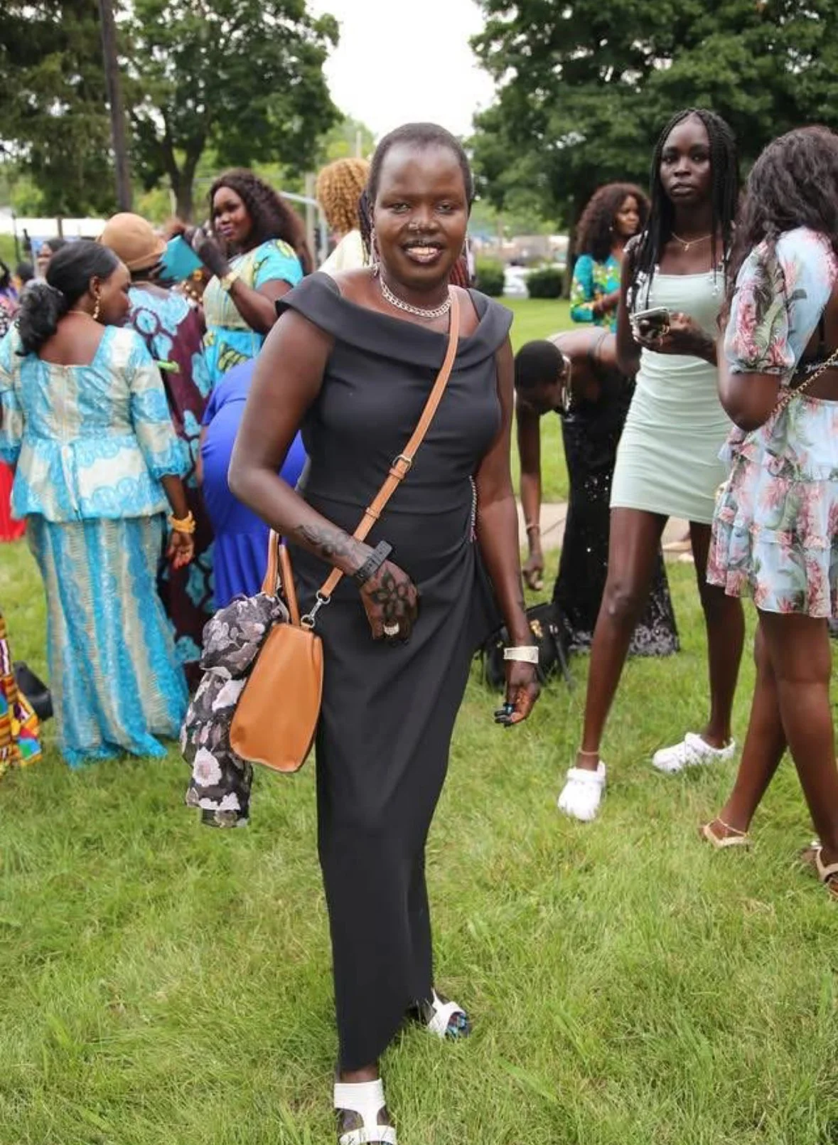 A woman in a black dress with jewelry and a brown shoulder bag standing on grass at an outdoor event with several other women dressed in colorful outfits in the background.