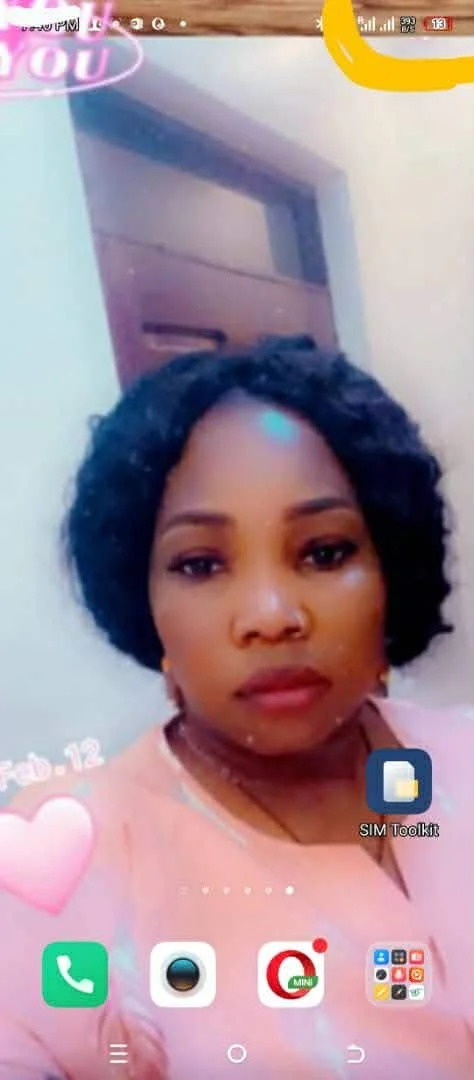 A woman with short curly black hair taking a selfie indoors. She is wearing a pink top and has a neutral expression. The phone screen shows icons for messaging, camera, and a folder labeled 'SIM Toolkit'.