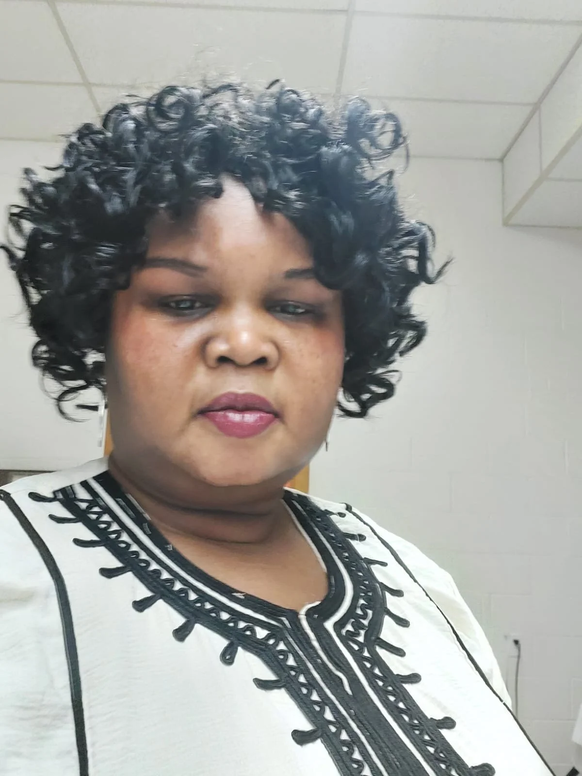A woman with short, black curly hair, wearing a white top with black embroidery, standing indoors against a plain background.