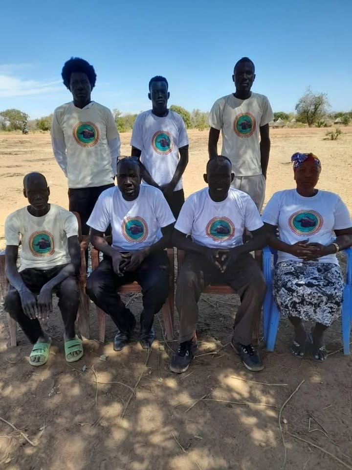 Seven people, four men and three women, are posing outdoors in a dry, rural area under a blue sky. They are wearing matching white T-shirts with a colorful circular logo featuring a green, purple, and blue design.