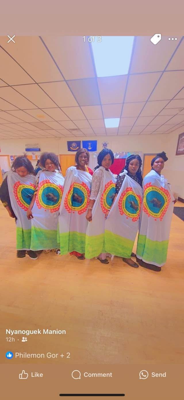 Six women standing in a row indoors, each wearing a white garment with a colorful emblem or logo on the front.