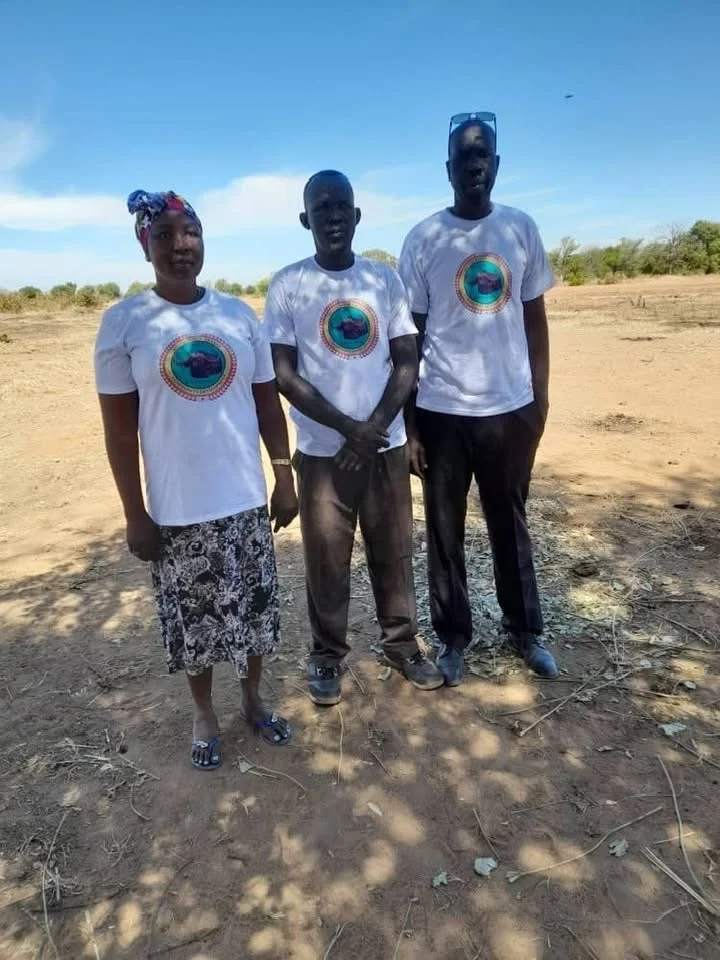 Three people standing outdoors in a dry, open area with a blue sky. All are wearing white T-shirts with a circular logo that appears to depict a car and text. The woman on the left is wearing a headscarf and patterned skirt, the man in the middle is shirtless, and the man on the right has glasses resting on his head and is dressed in dark pants.