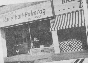 Black and white photo of a storefront with the sign 'Kane Hall-Palmtag Insurance' and a striped awning.