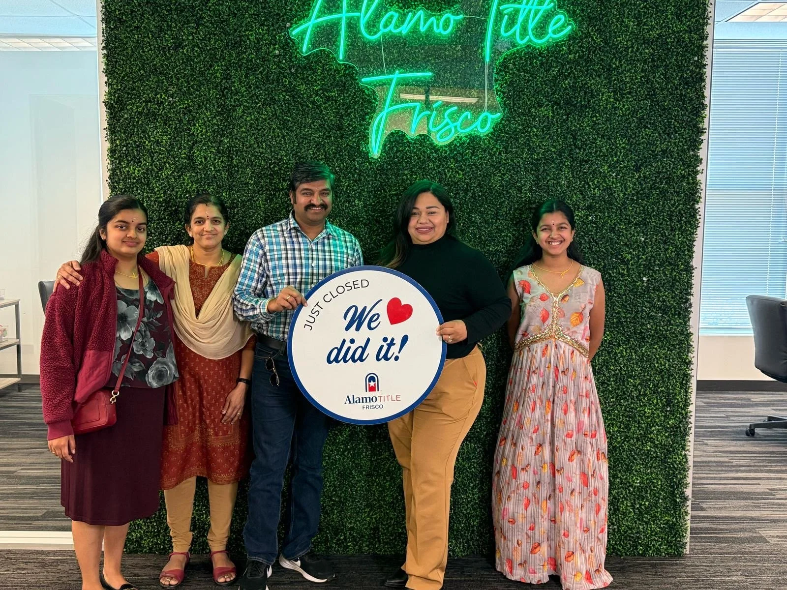 Group of five people standing in front of a green wall with a neon sign that reads, 'Alamo Title Frisco'. They are holding a circular sign that says, 'Just closed, We love did it!' The group is smiling and celebrating an achievement.