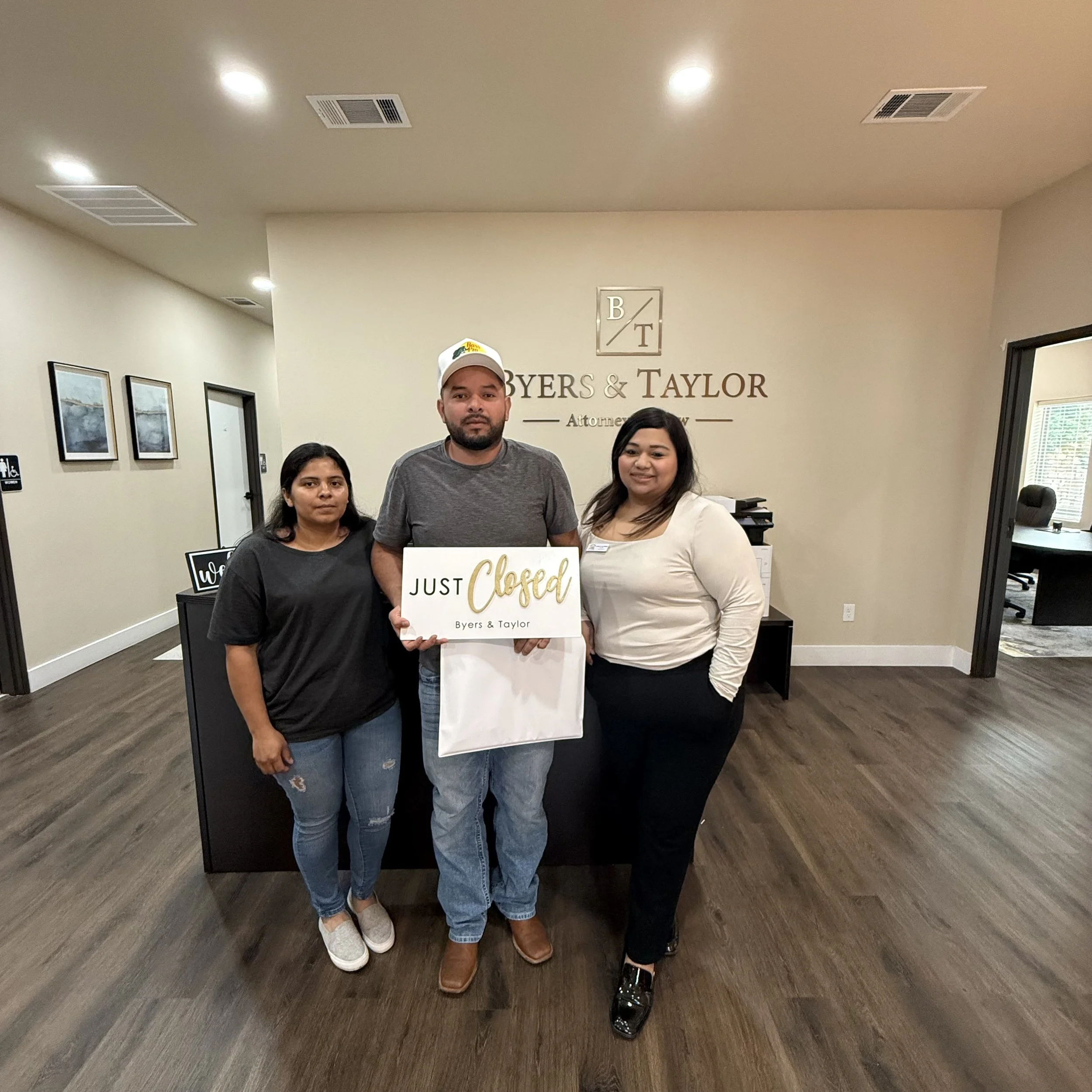 Three people standing in an office lobby, holding a sign that says "Just Closed" with the law firm's name "Byers & Taylor" on it. The man in the middle has a beard, wears a gray t-shirt, jeans, brown shoes, and a white cap. The woman on the right is dressed in a white blouse and black pants. The woman on the left is in a black top and ripped jeans.