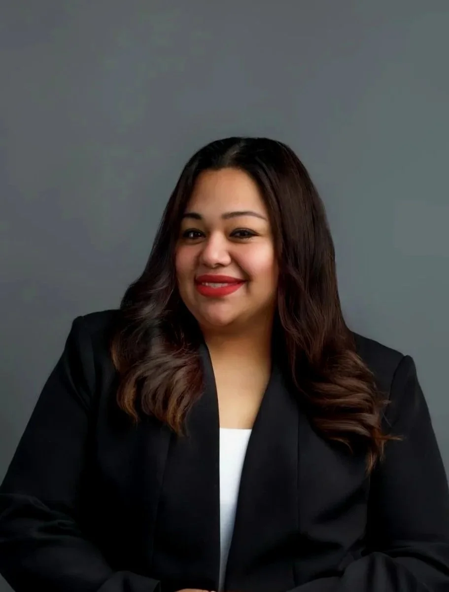 A woman with long wavy dark hair, wearing a black blazer over a white top, smiling against a plain gray background top producing professional