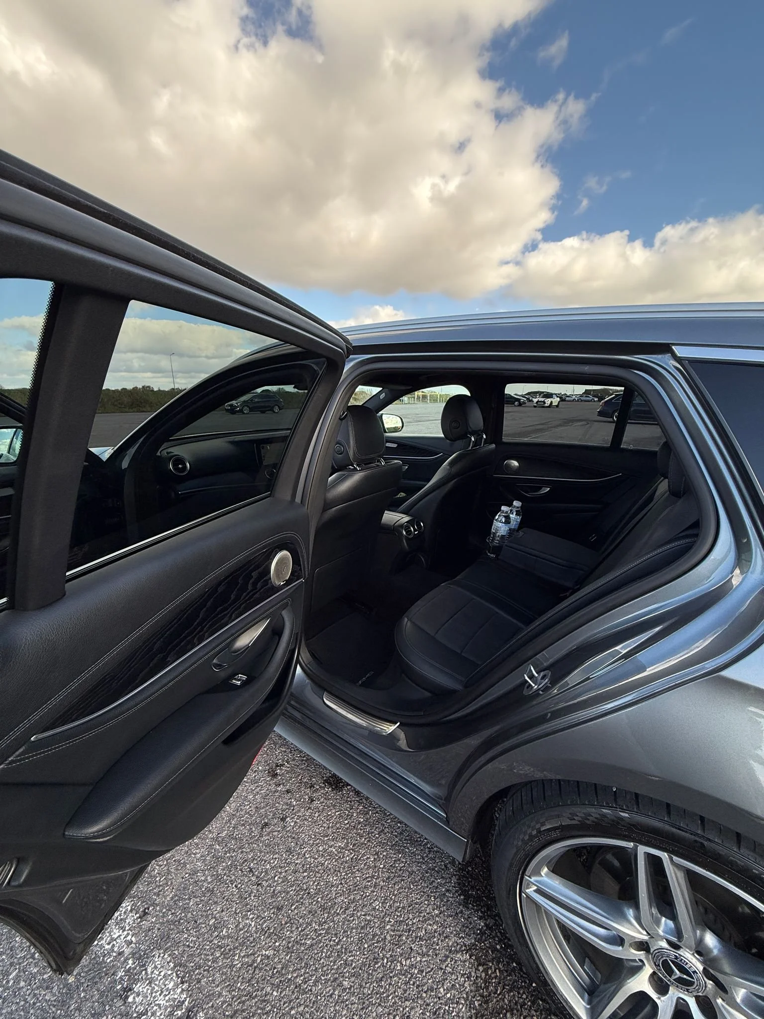 Open car door revealing black leather interior, parked in a lot with other vehicles and a cloudy sky above.