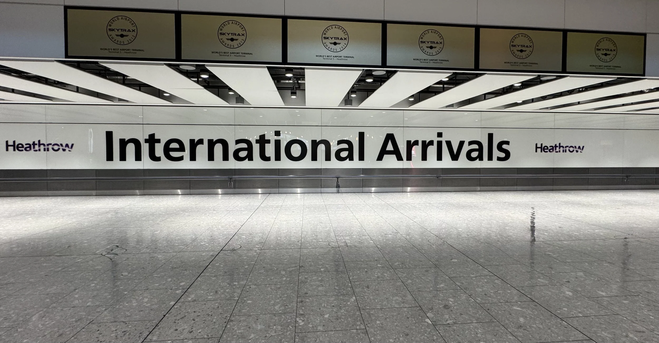 View of Heathrow Airport's International Arrivals entrance with large black and purple signage on reflective glass wall, tiled floor, and ceiling with light fixtures.