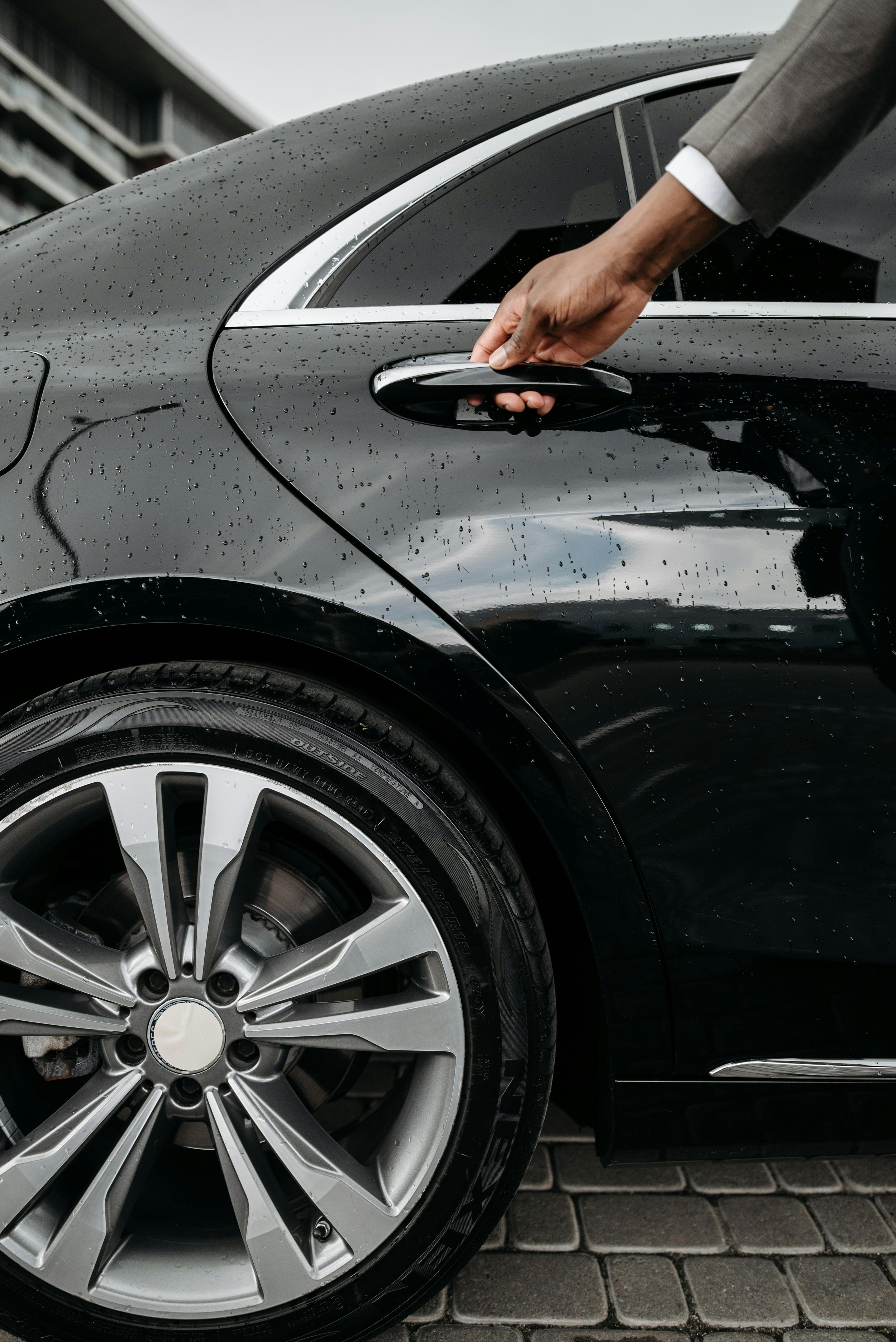 Close-up of a black luxury car's door handle being opened by a man's hand. The car has raindrops on its surface and is parked on a cobblestone street.