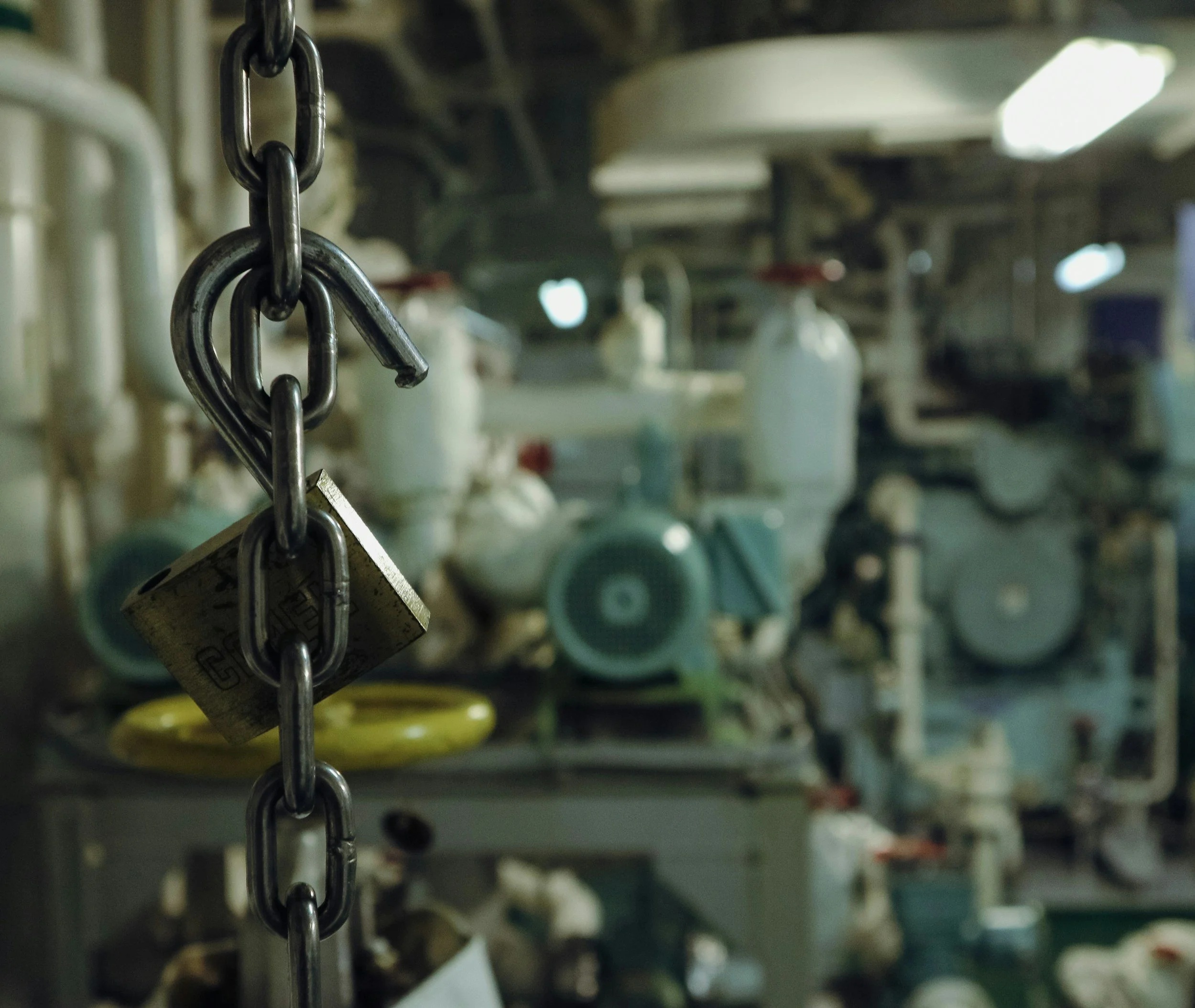 A chain with a padlock hanging from it in the foreground, with industrial equipment and machinery blurred in the background.
