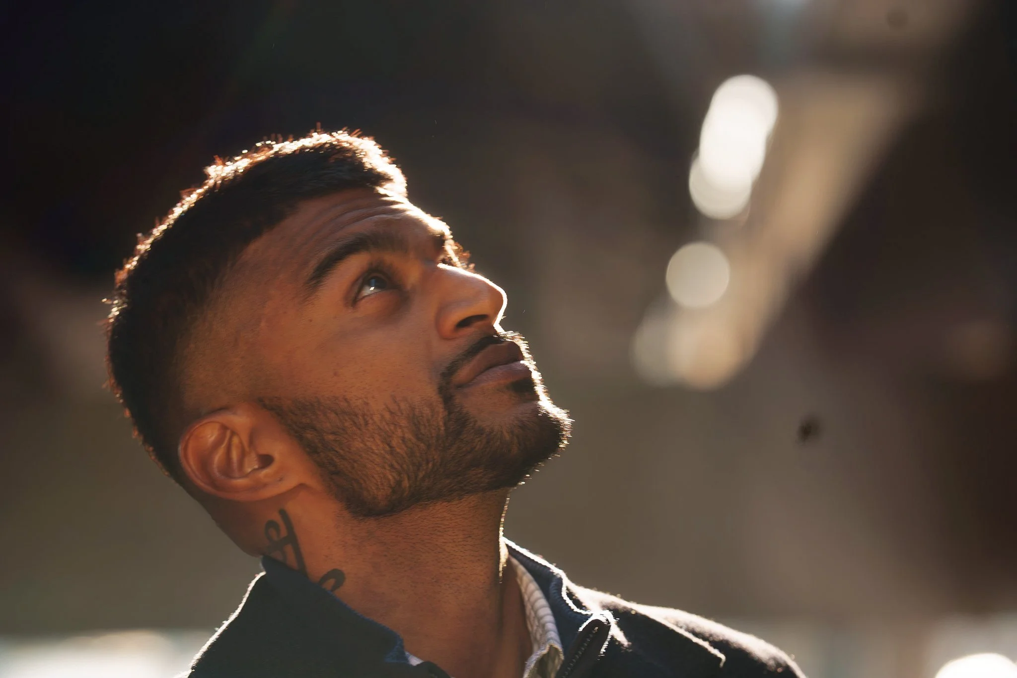 A man with a short beard and tattoos on his neck, looking upward. He has short, dark hair and is wearing a dark jacket with a white shirt underneath. The background is blurred with bright light spots.