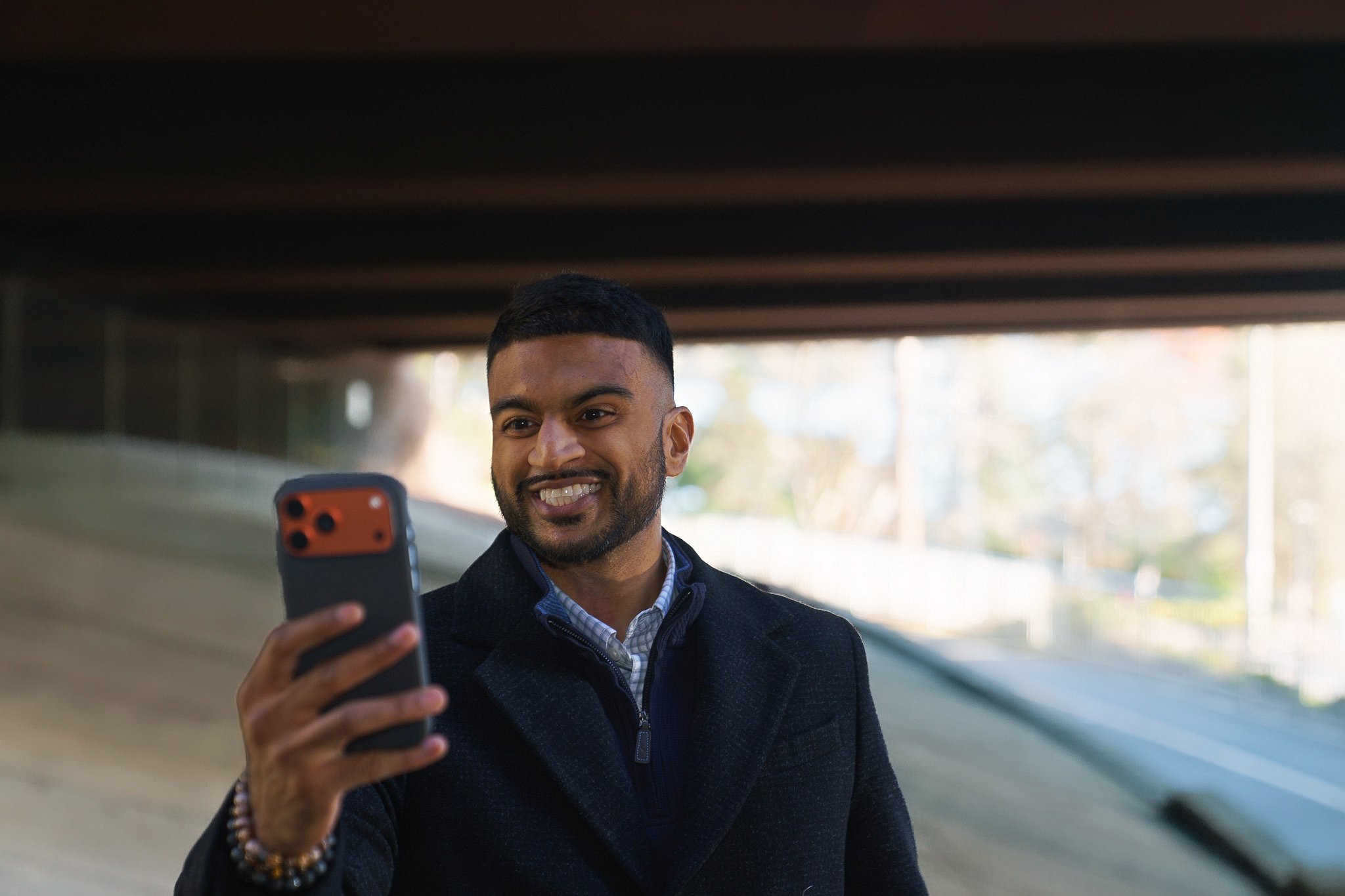 A man smiling and taking a selfie with a phone under a bridge or overpass.