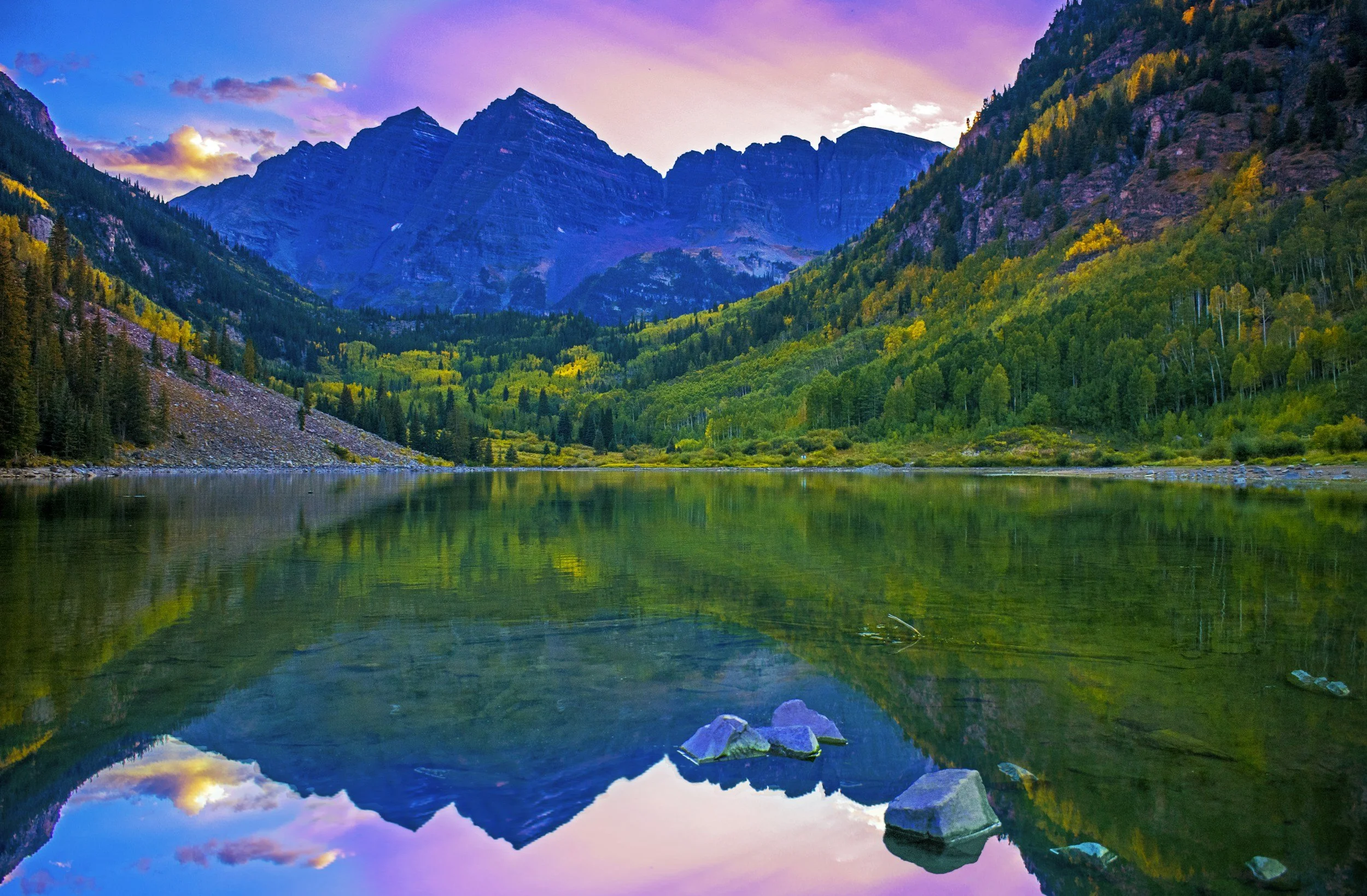 Scenic view of a lake surrounded by lush green forests and towering mountains under a vibrant pink and purple sky.
