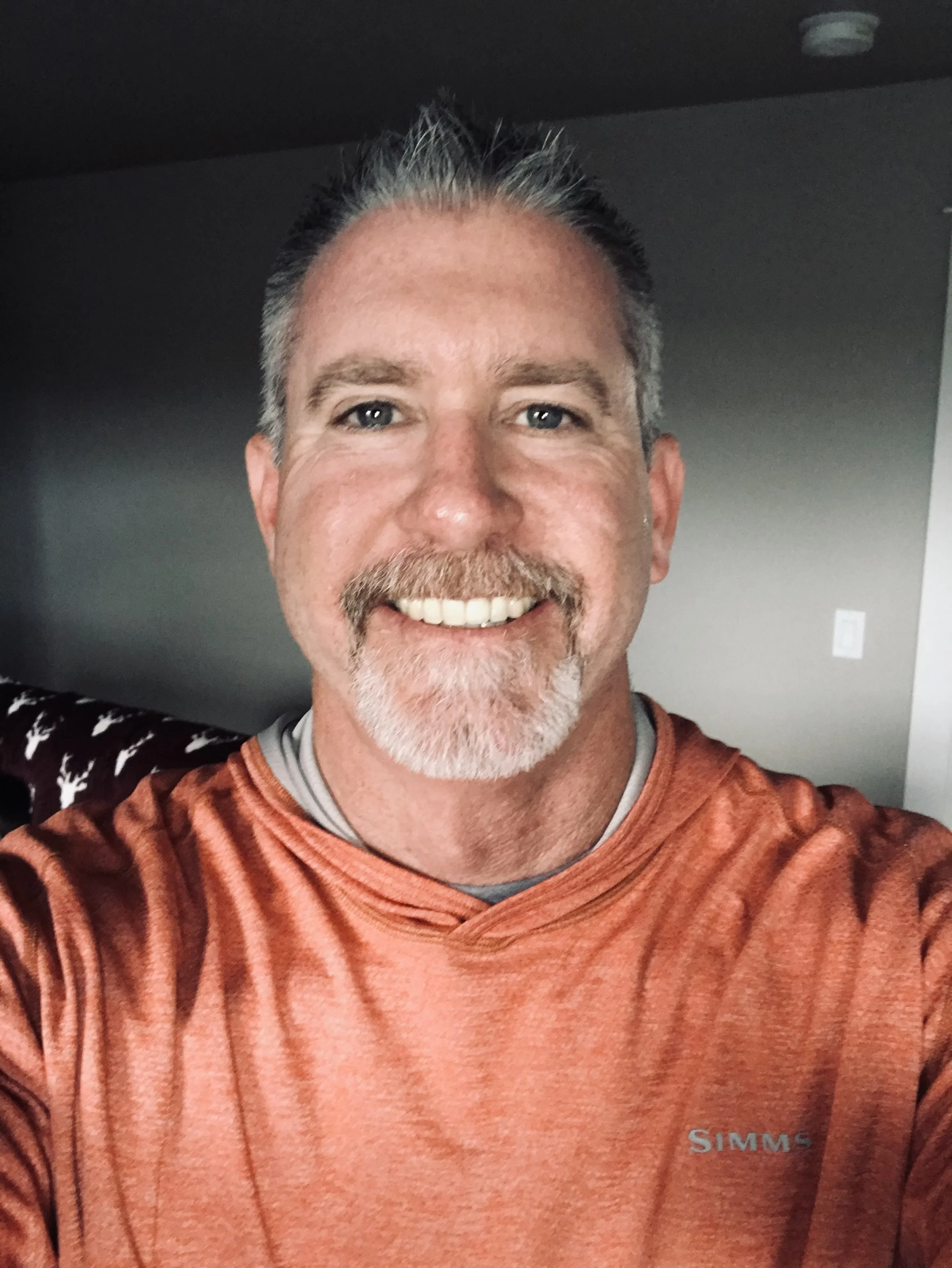 A smiling middle-aged man with gray hair, a beard, and blue eyes, wearing an orange athletic shirt, taking a selfie indoors.