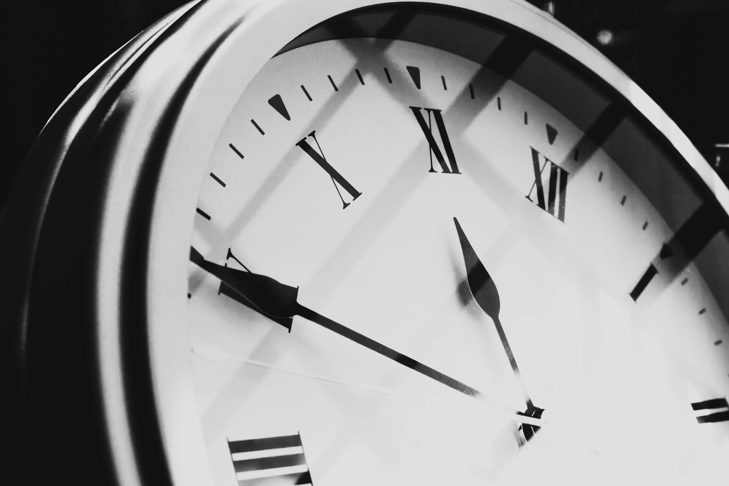 Close-up of a round analog clock showing nearly 10:54, with a reflection on the glass cover and a black-and-white color scheme.