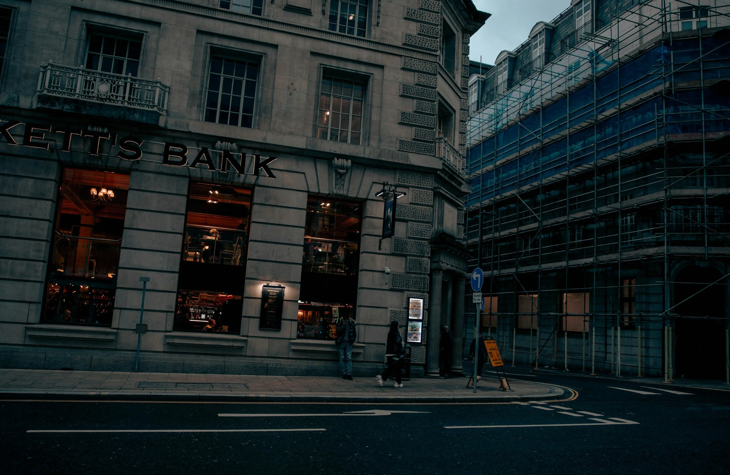 Street view of a retail building with a bank on the ground floor, surrounded by scaffolding on one side, during evening or dusk.