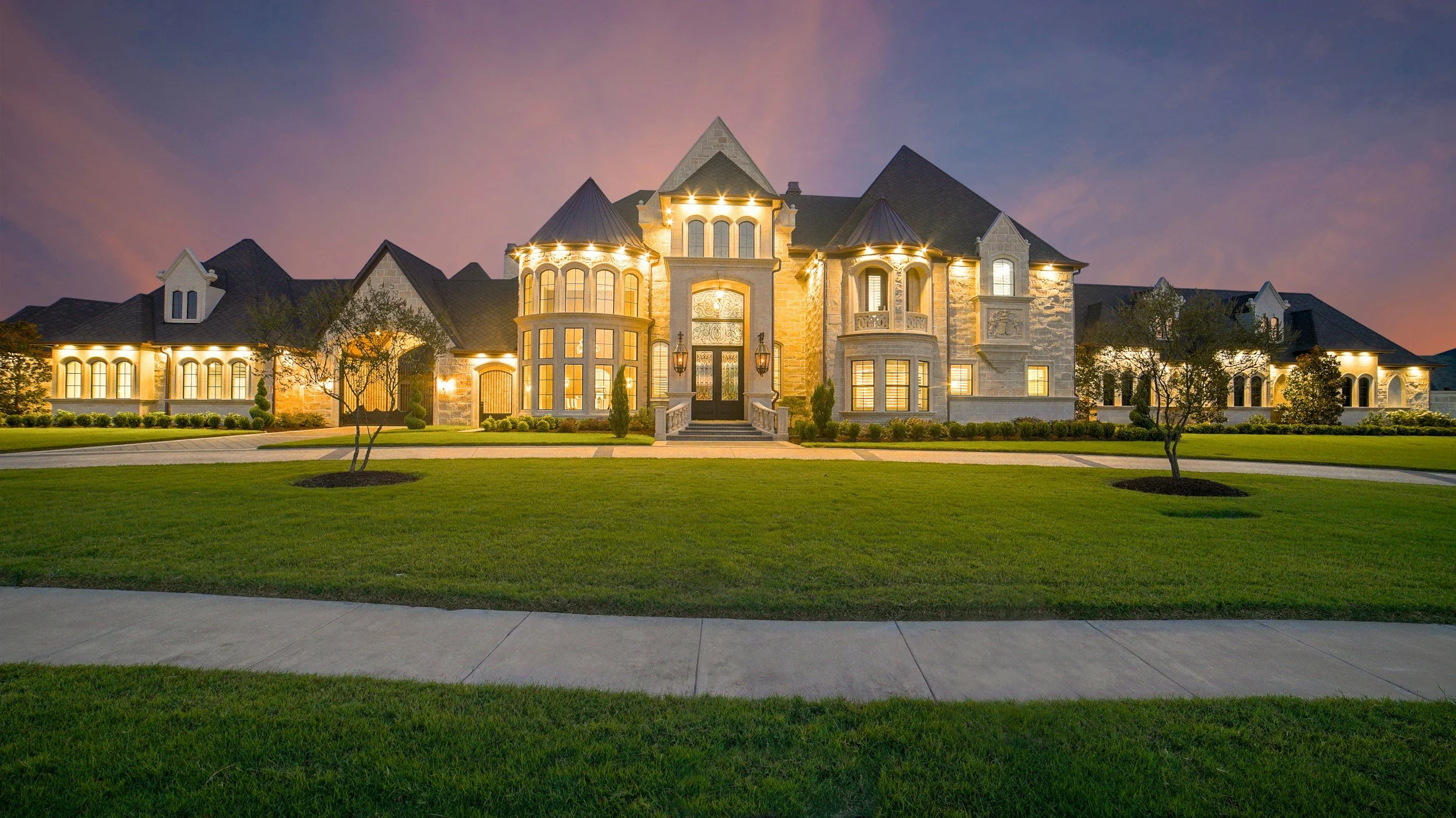 Large, elegant mansion with stone facade, multiple turrets, and illuminated windows at dusk, surrounded by a manicured lawn and trees.